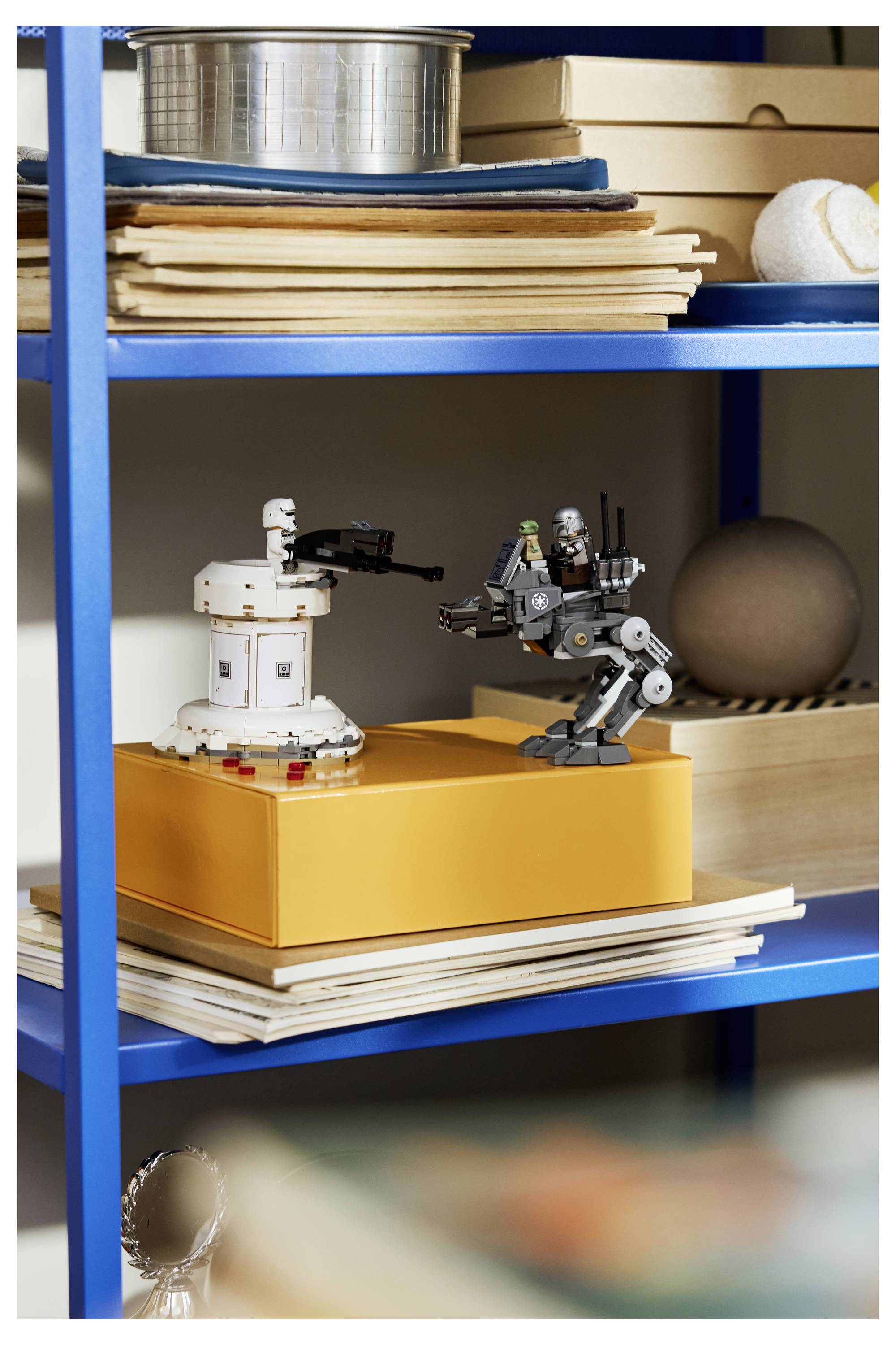 Ein blaues Regal hält gestapelte Blätter Papier und kleine Gegenstände, einschließlich zwei robotähnlicher Figuren, eine graue und eine weiße, auf einer orangefarbenen Schachtel.