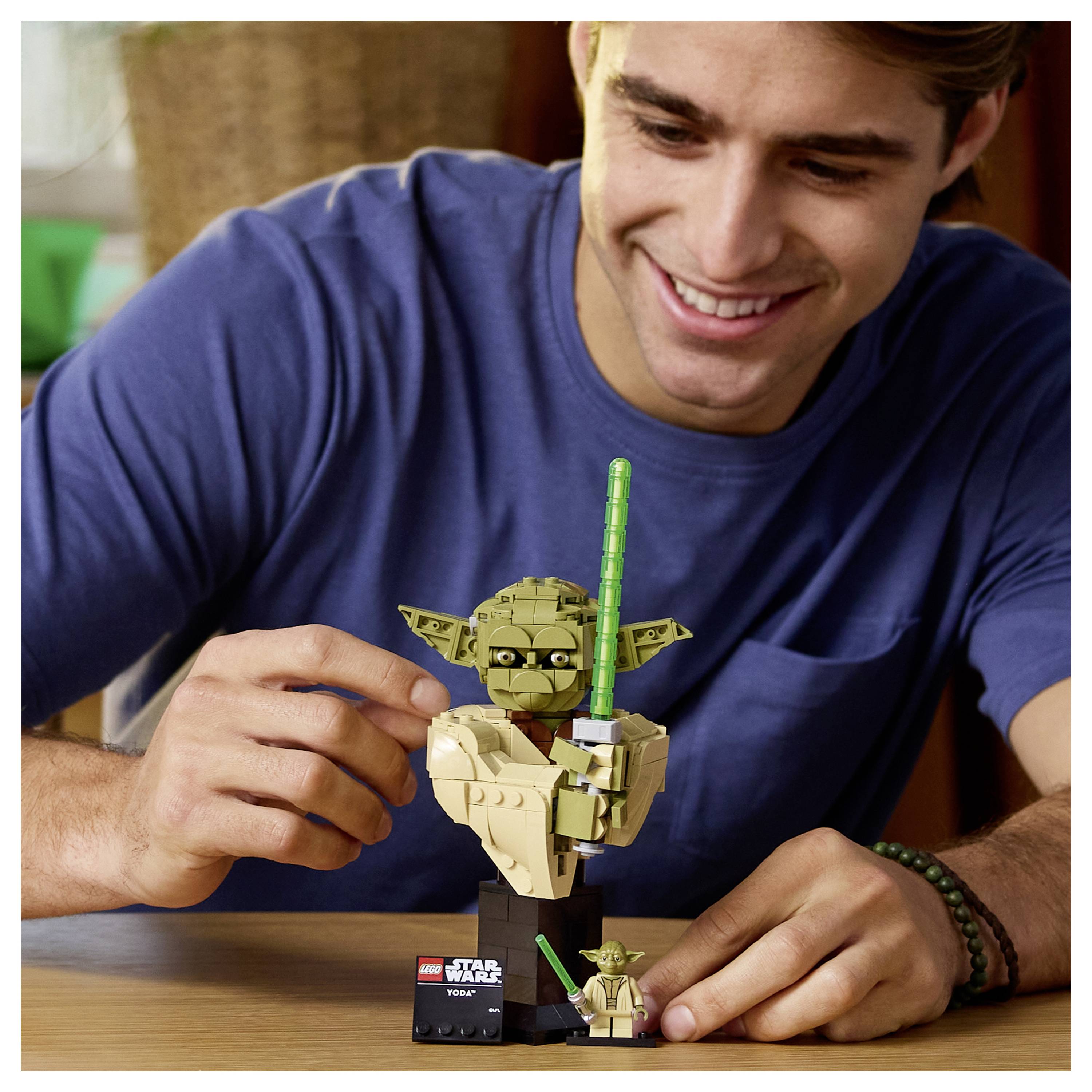 Eine Person, die lächelt, während sie ein Star Wars Yoda LEGO-Set auf einem Tisch zusammenbaut, ein Stück in der Hand haltend, mit einer Miniaturfigur daneben.
