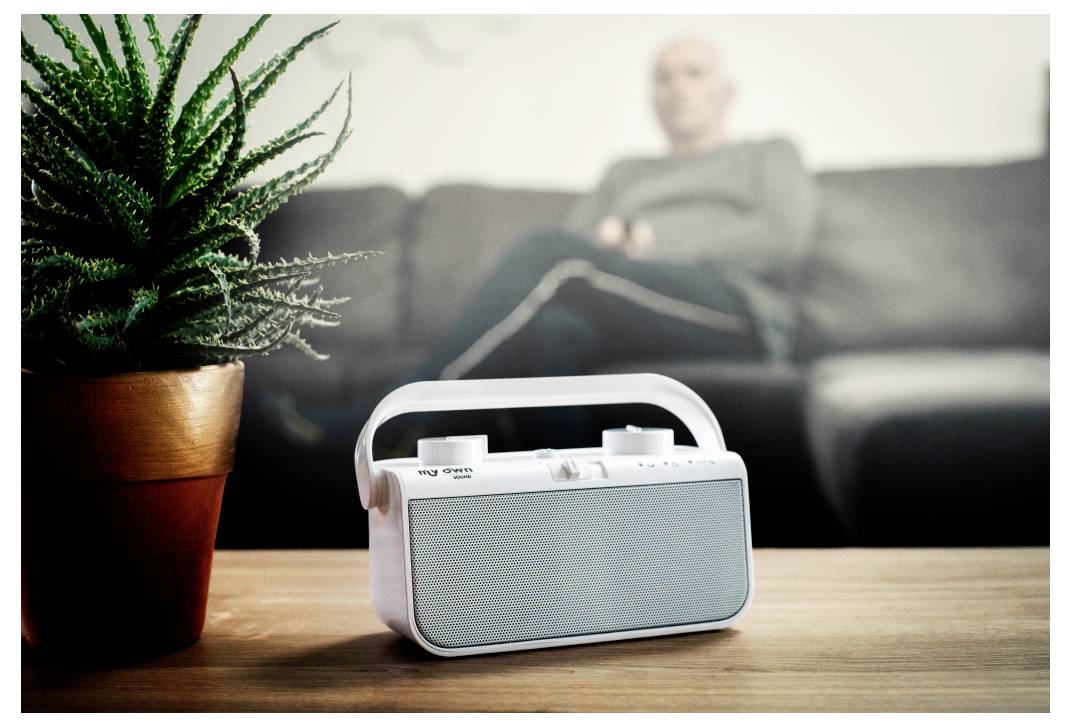 Ein kleines weißes Radio auf einem Holztisch, eine Topfpflanze daneben; eine Person sitzt verschwommen im Hintergrund auf einem Sofa.