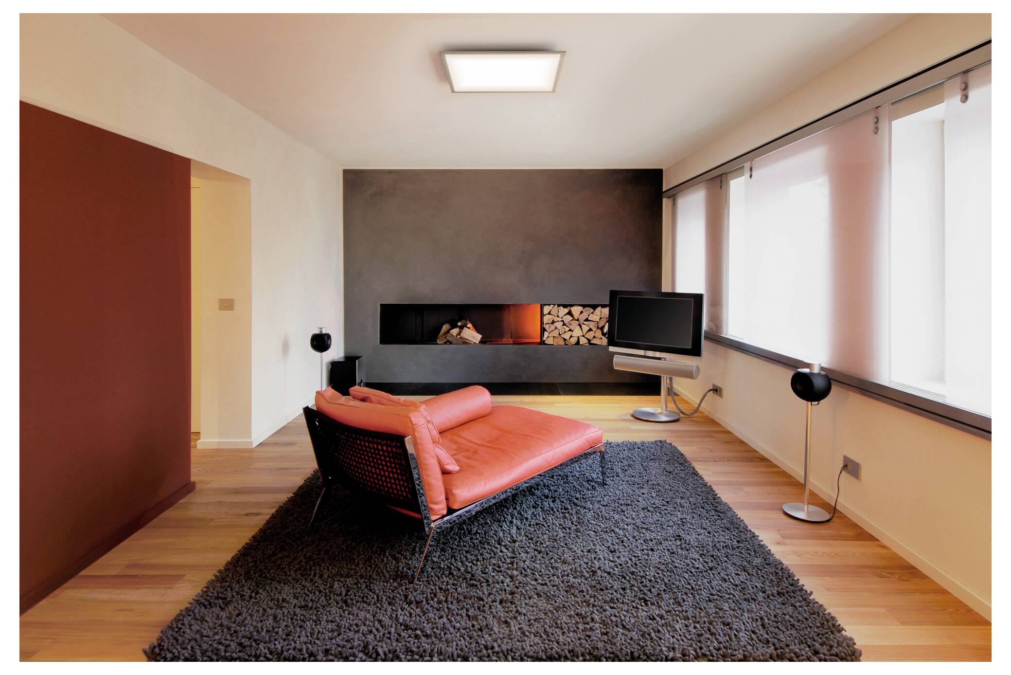 Ein modernes Wohnzimmer mit einer roten Récamiere auf einem grauen Teppich, einem an der Wand montierten Fernseher und einem Kamin mit gestapelten Holzscheiten, unter sanfter Beleuchtung.