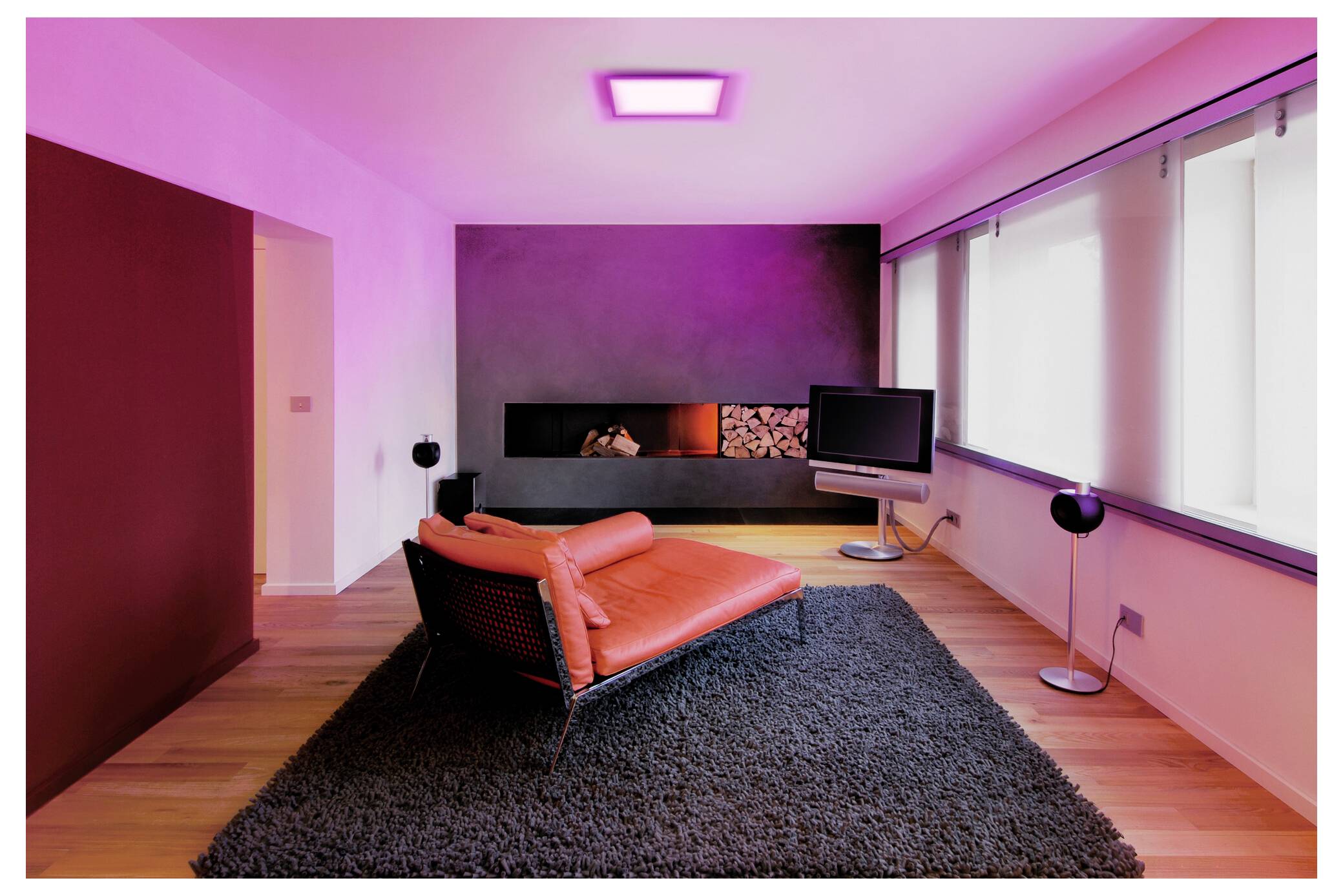 Ein modernes Wohnzimmer mit einer roten Récamiere, schwarzem Teppich, wandmontiertem Fernseher und Kamin. Der Raum wird von rosa Beleuchtung durchflutet.