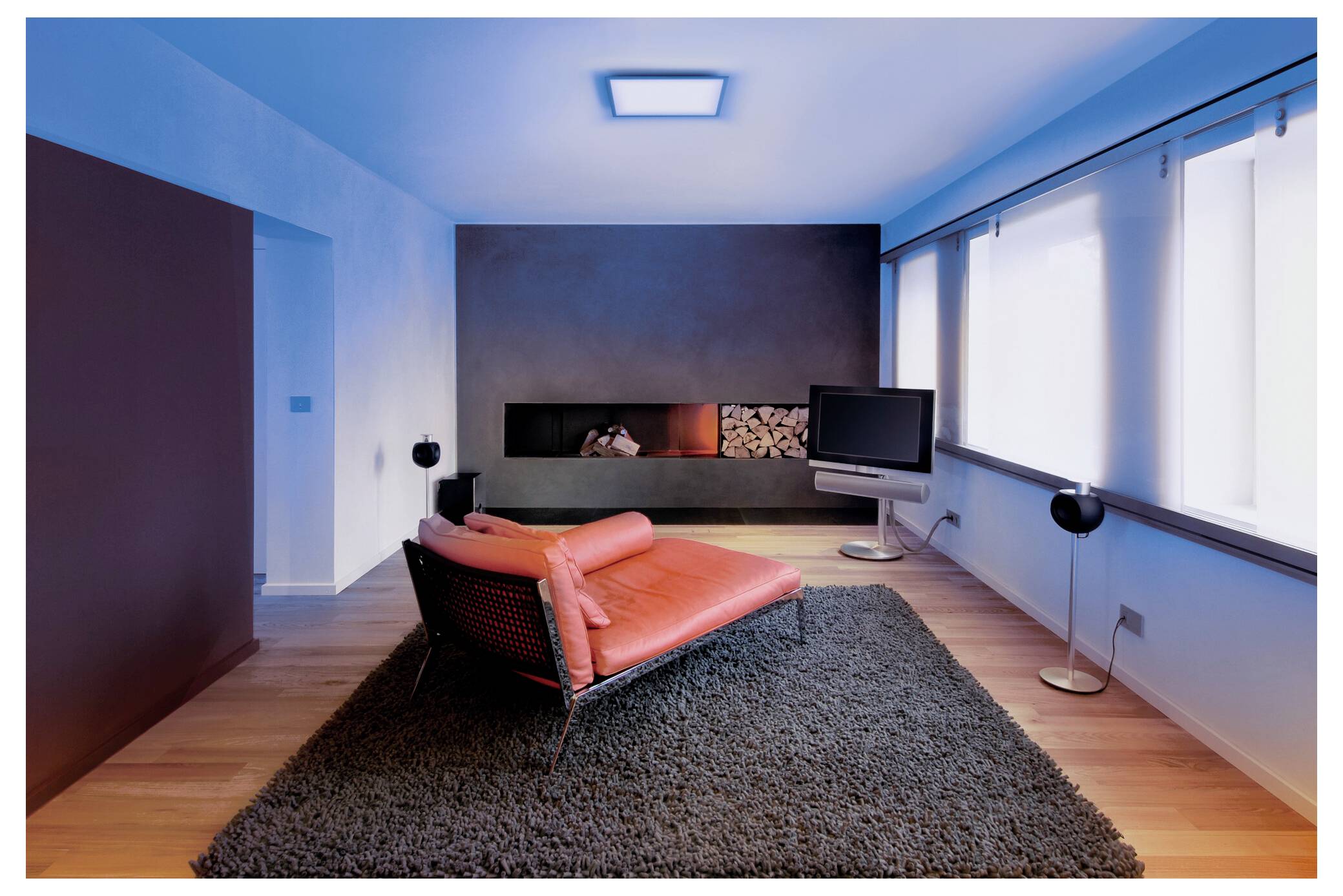 Ein modernes Wohnzimmer mit einem roten Loungesessel auf einem grauen Teppich, einem Flachbild-Fernseher und einem Kamin mit Holzscheiten. Große Fenster lassen natürliches Licht herein.