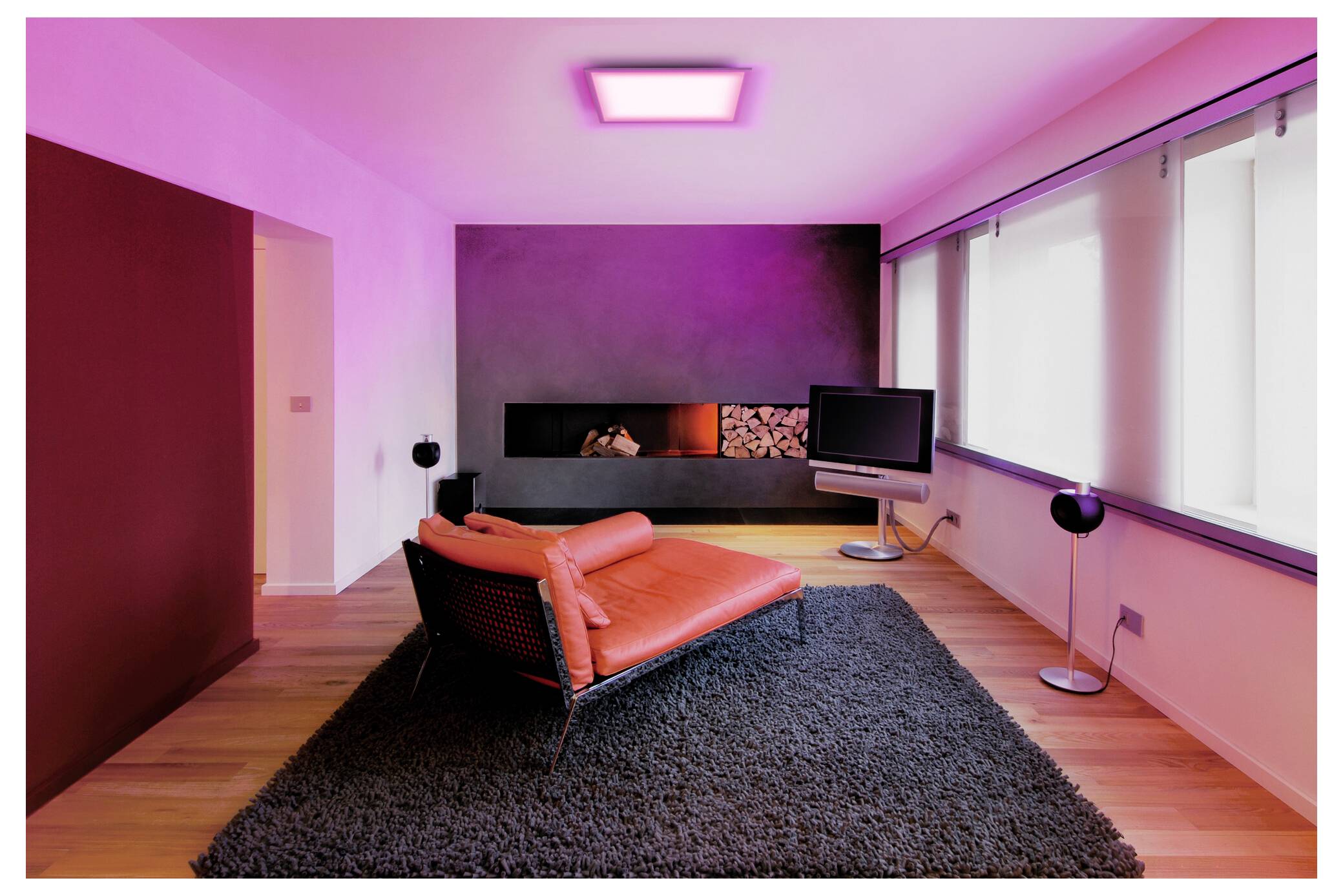 Ein modernes Wohnzimmer mit einem roten Stuhl auf einem grauen Teppich, violetter Beleuchtung, einer wandmontierten Fernseher und einem eingebauten Kamin mit Holzlagerung.
