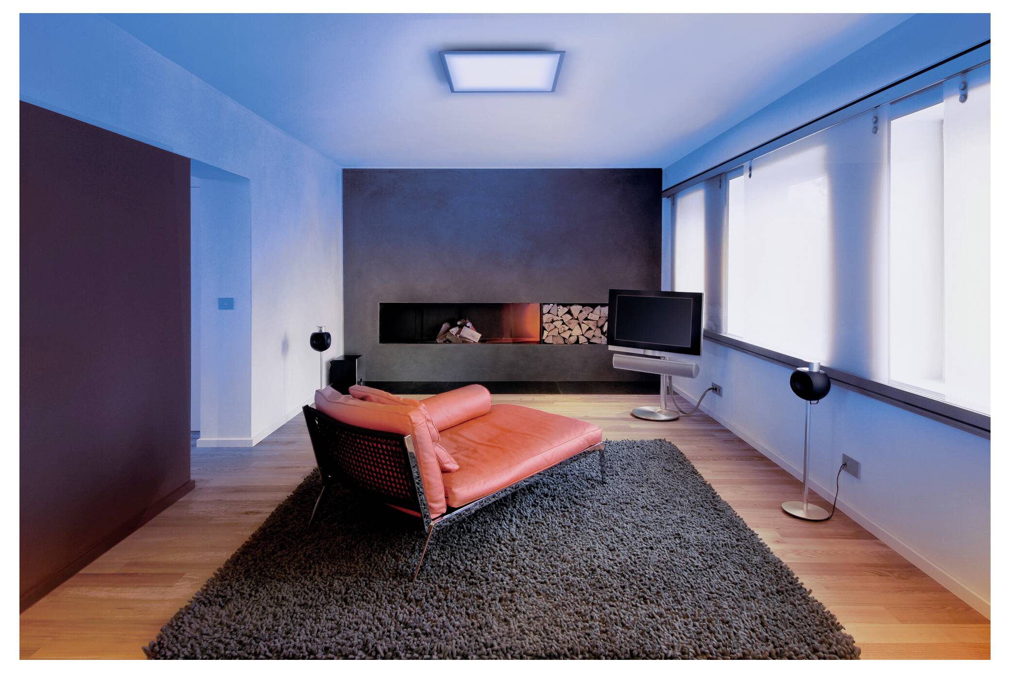 Ein modernes Wohnzimmer mit einer roten Récamiere auf einem dunklen Teppich, einem wandmontierten Kamin, einem Flachbild-Fernseher und großen Fenstern auf der rechten Seite.