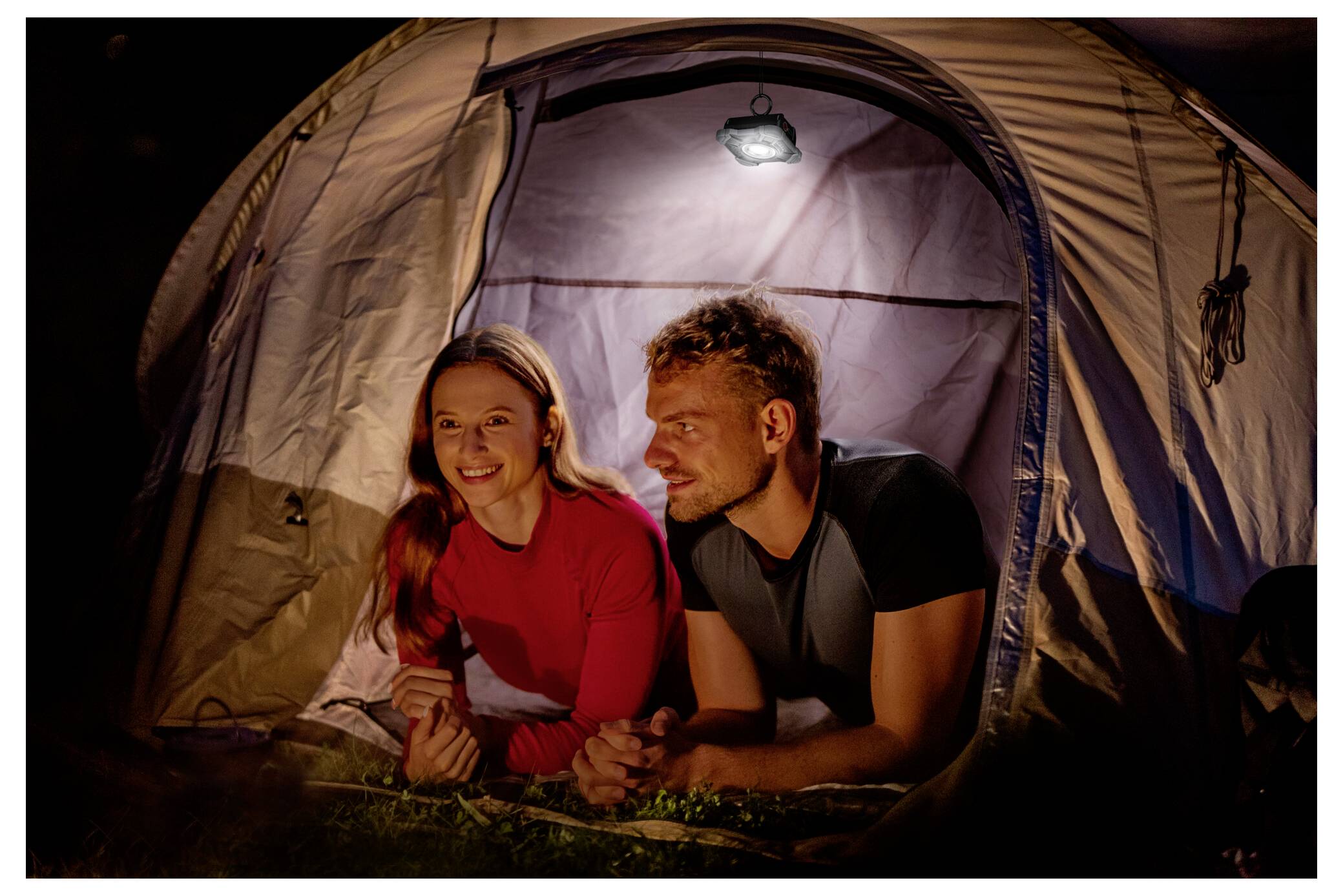 Ein Paar liegt nachts in einem beleuchteten Zelt, lächelt und spricht, während draußen Campingausrüstung zu sehen ist. Die Szene deutet auf ein entspannendes Outdoor-Abenteuer hin.