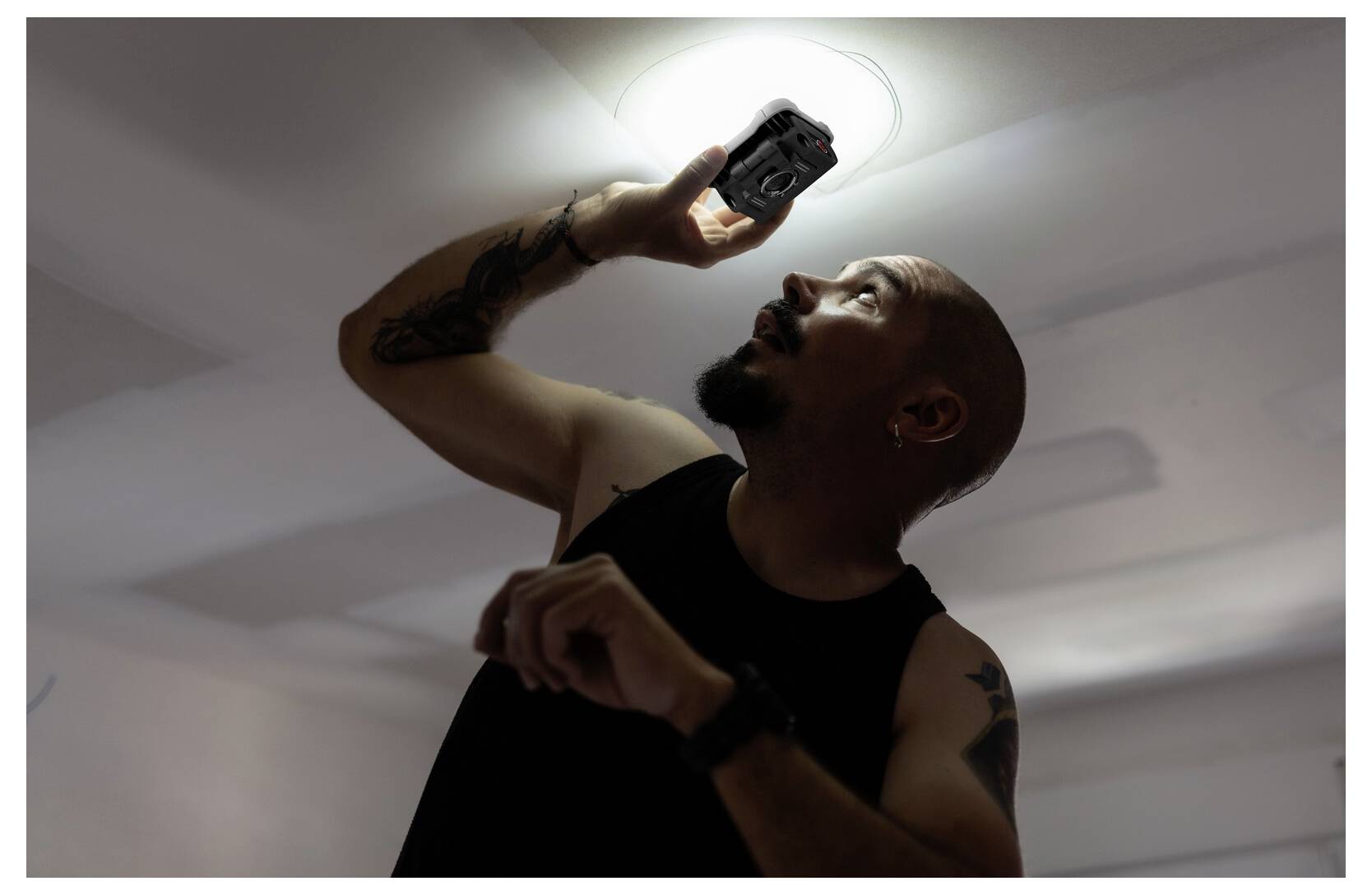 Eine Person steht auf einer Leiter, hält eine Kamera nach oben zu einer beleuchteten Deckenlampe und inspiziert oder fotografiert etwas darüber.