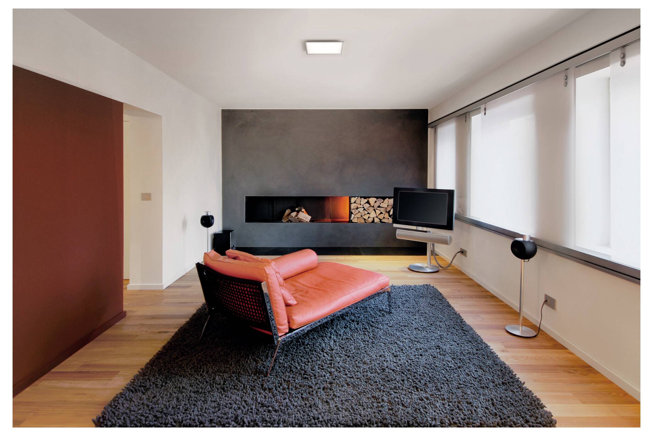 Ein modernes Wohnzimmer mit einer roten Récamiere auf einem grauen Teppich, einer schwarzen Akzentwand mit Brennholz und einem großen Flachbildfernseher auf einem Ständer.