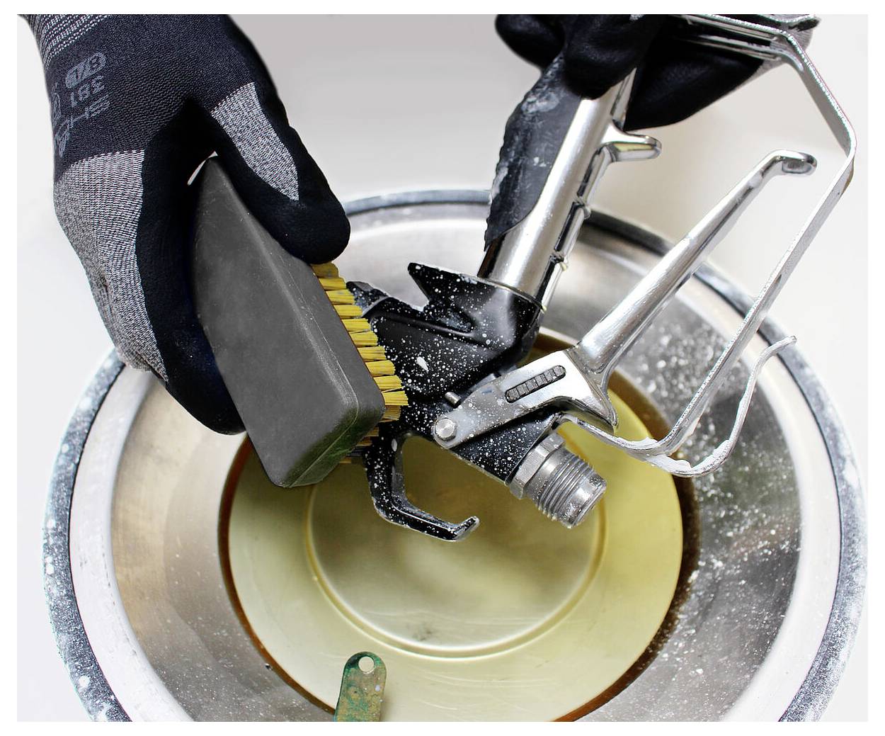 Eine Person mit Handschuhen verwendet einen kleinen Pinsel, um eine Sprühpistole über einem Behälter zu reinigen und damit die Wartung von Malerausrüstung demonstriert.