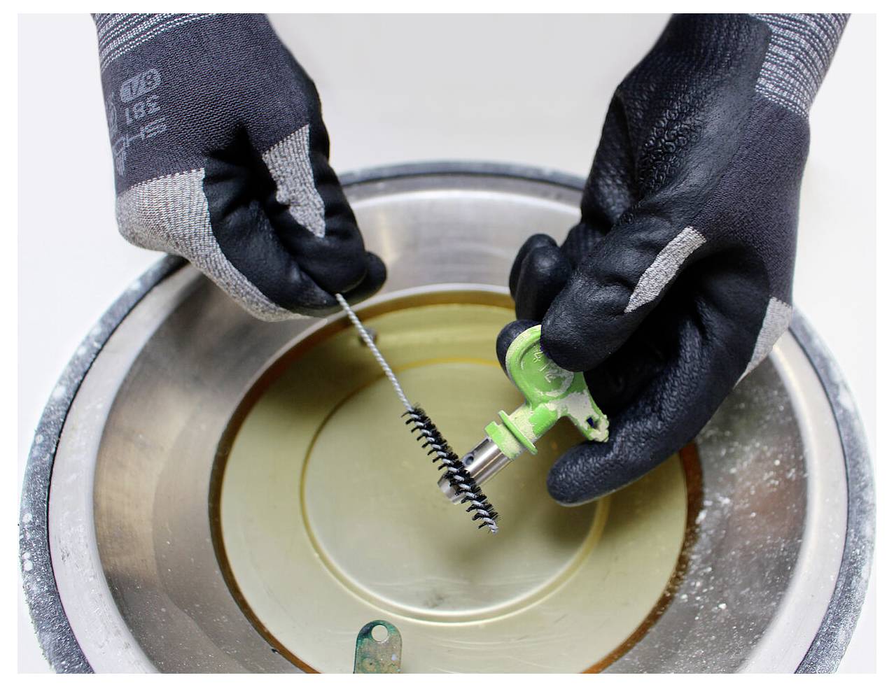 Zwei mit Handschuhen bekleidete Hände, die ein kleines Metallstück mit einer Drahtbürste über einen runden Metallbehälter reinigen.