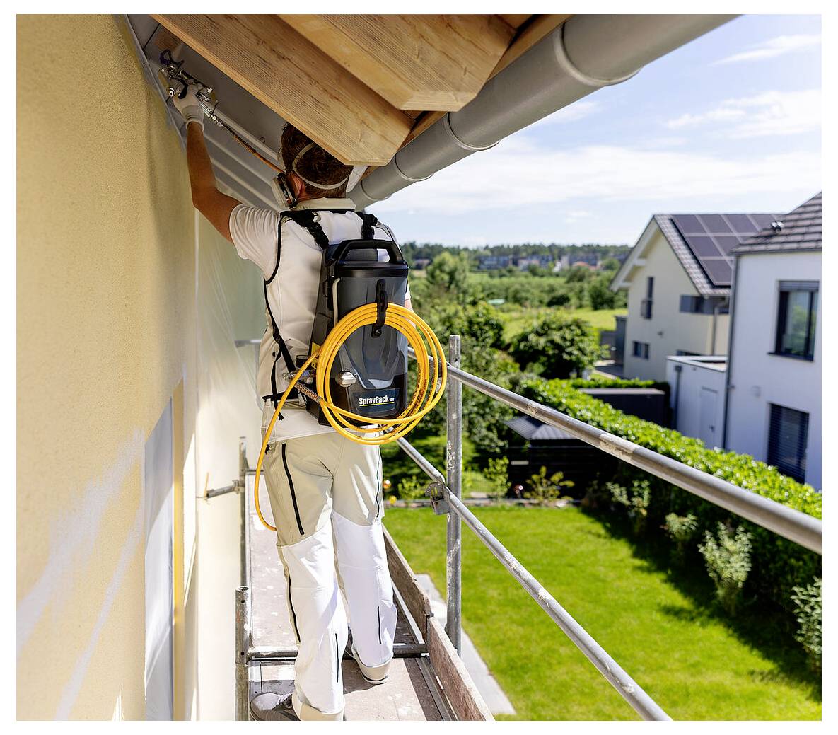 Eine Person steht auf Gerüsten und bemalt die Außenseite eines Hauses mit einer Sprühmaschine. Im Hintergrund sind Häuser und Vegetation zu sehen.