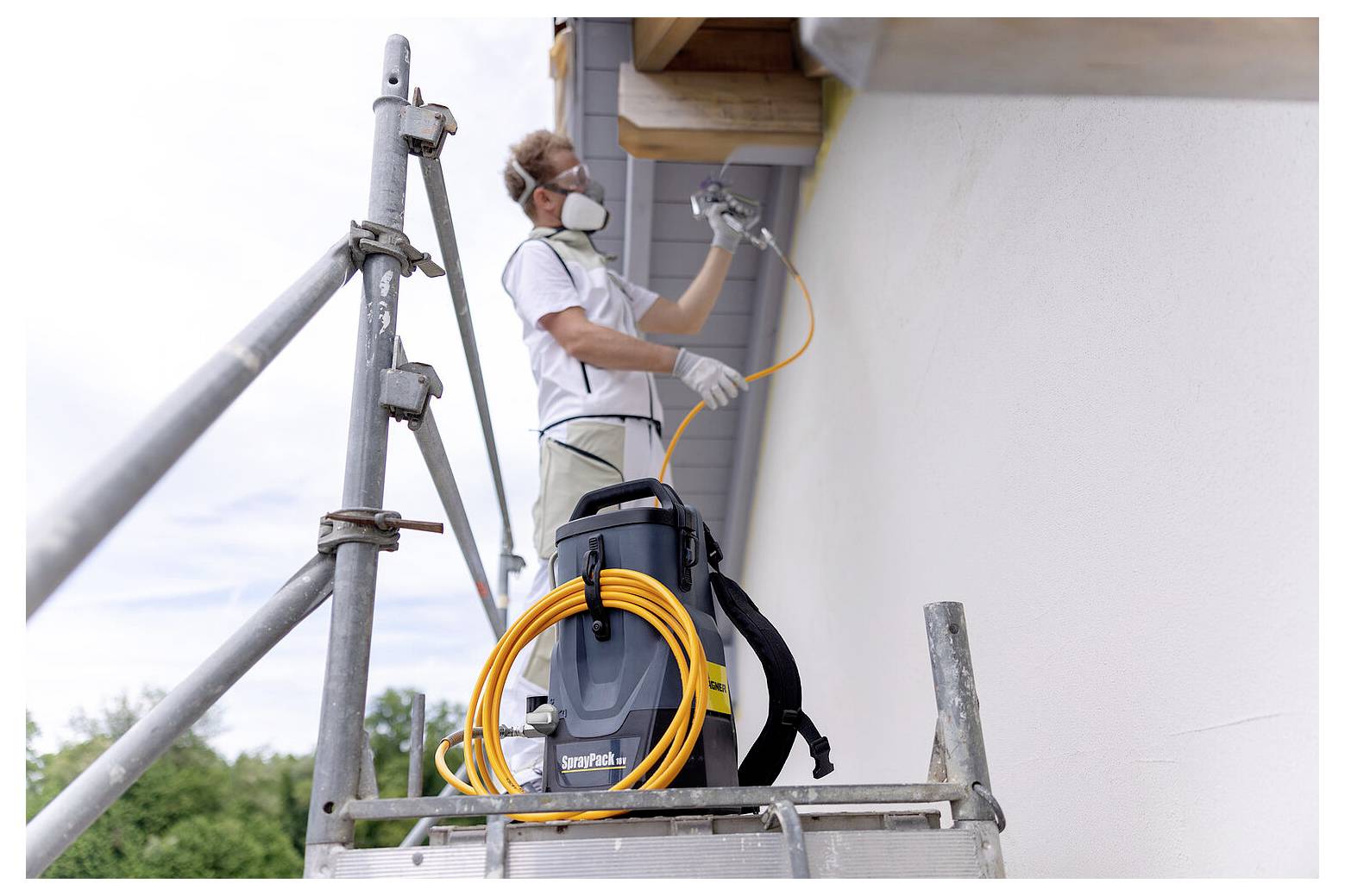Eine Person mit Maske und Handschuhen verwendet einen Farbsprüher an der Außenseite eines Hauses, während sie auf Gerüsten steht. Ein Schlauch und weitere Ausrüstung befinden sich in der Nähe.