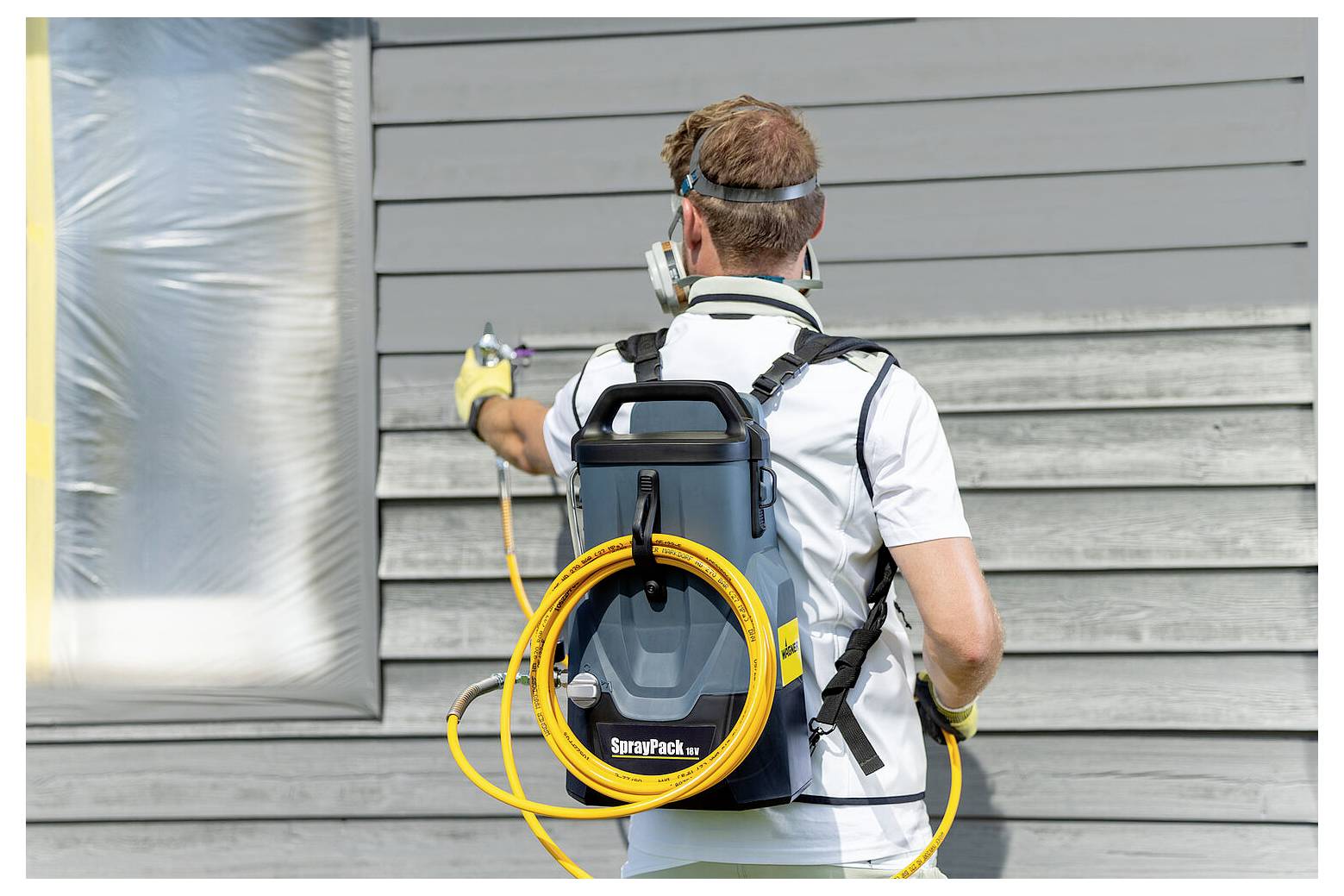 Eine Person mit einem Rückentragsystem für Sprühfarbe bemalt die Außenwand eines Hauses mit grauer Verschalung. Die Person steht mit dem Gesicht zur Wand.