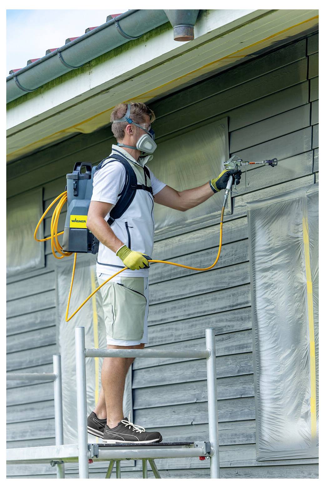 Eine Person in Sicherheitsausrüstung verwendet eine Spritzpistole, um die Seite eines Hauses zu streichen, während sie auf Gerüsten steht. Der Bereich ist zum Schutz abgedeckt.