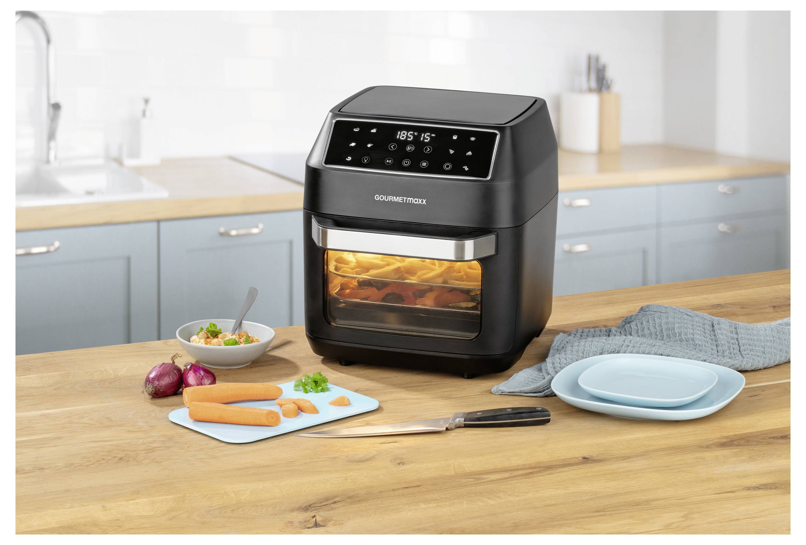 Ein schwarzer digitaler Heißluftfritteur auf einer Küchenarbeitsplatte brät Hähnchen. In der Nähe befinden sich gehackte Gemüsestücke, ein Schneidebrett mit Karotten, ein Messer und blaue Teller.