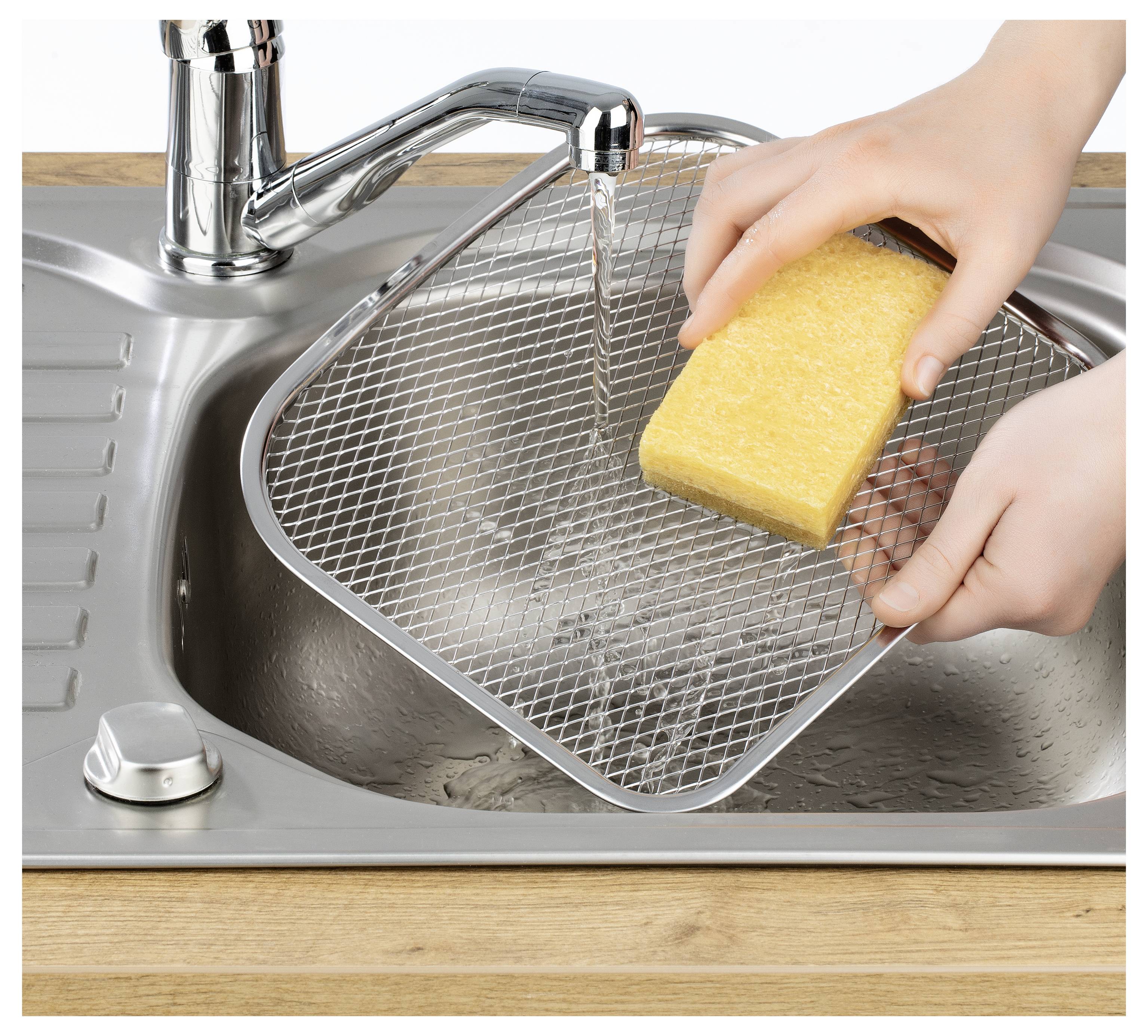 Eine Person reinigt ein Metallgitter mit einem gelben Schwamm unter fließendem Wasser in einem Küchenbecken.