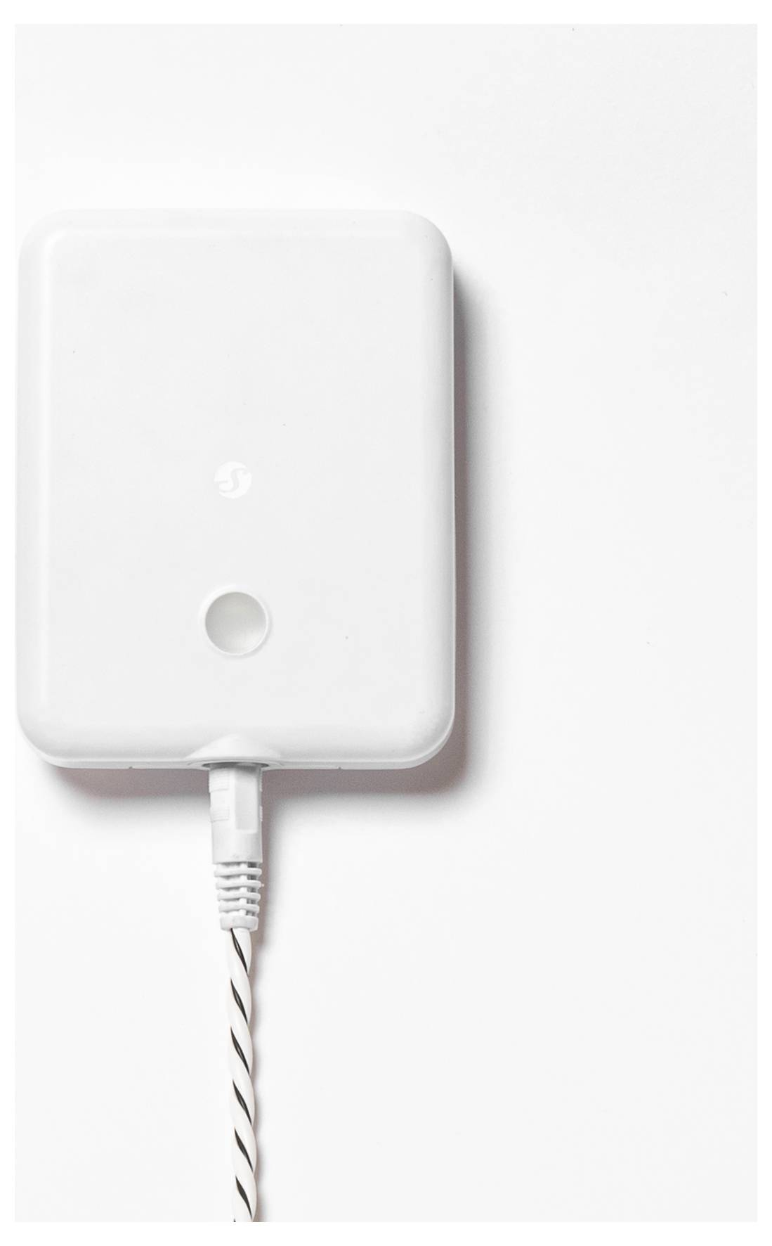 Weißes rechteckiges elektrisches Gerät mit einer kreisförmigen Taste, das durch ein weißes Kabel verbunden ist, vor einem einfarbig weißen Hintergrund.