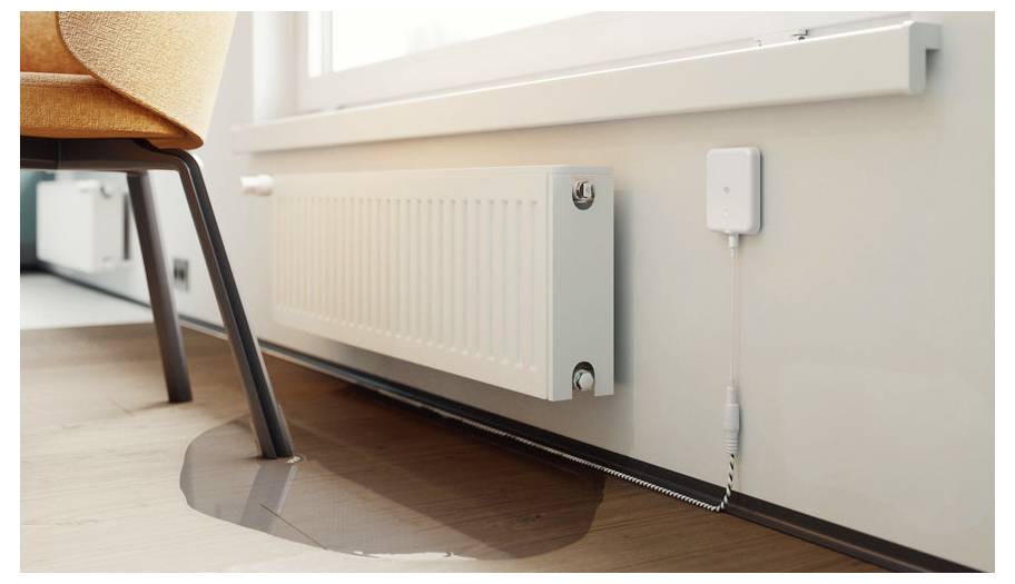 Eine kleine Wasserpfütze auf einem Holzboden deutet auf einen Heizkörperleck hin; ein weißer Sensor ist in der Nähe zur Überwachung angebracht.