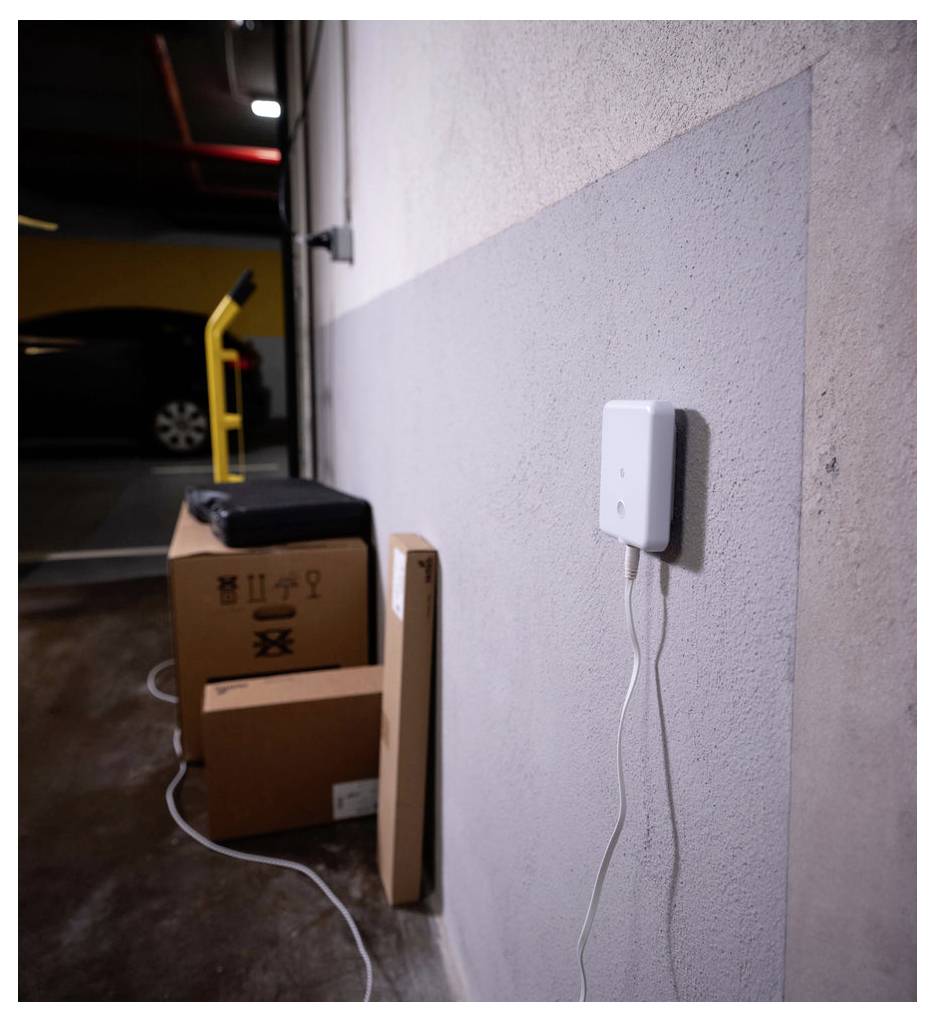 Kisten und ein schwarzer Koffer sind an einer Wand eines Parkhauses gestapelt, wobei ein Gerät an der Wand montiert ist und durch ein weißes Kabel verbunden ist.