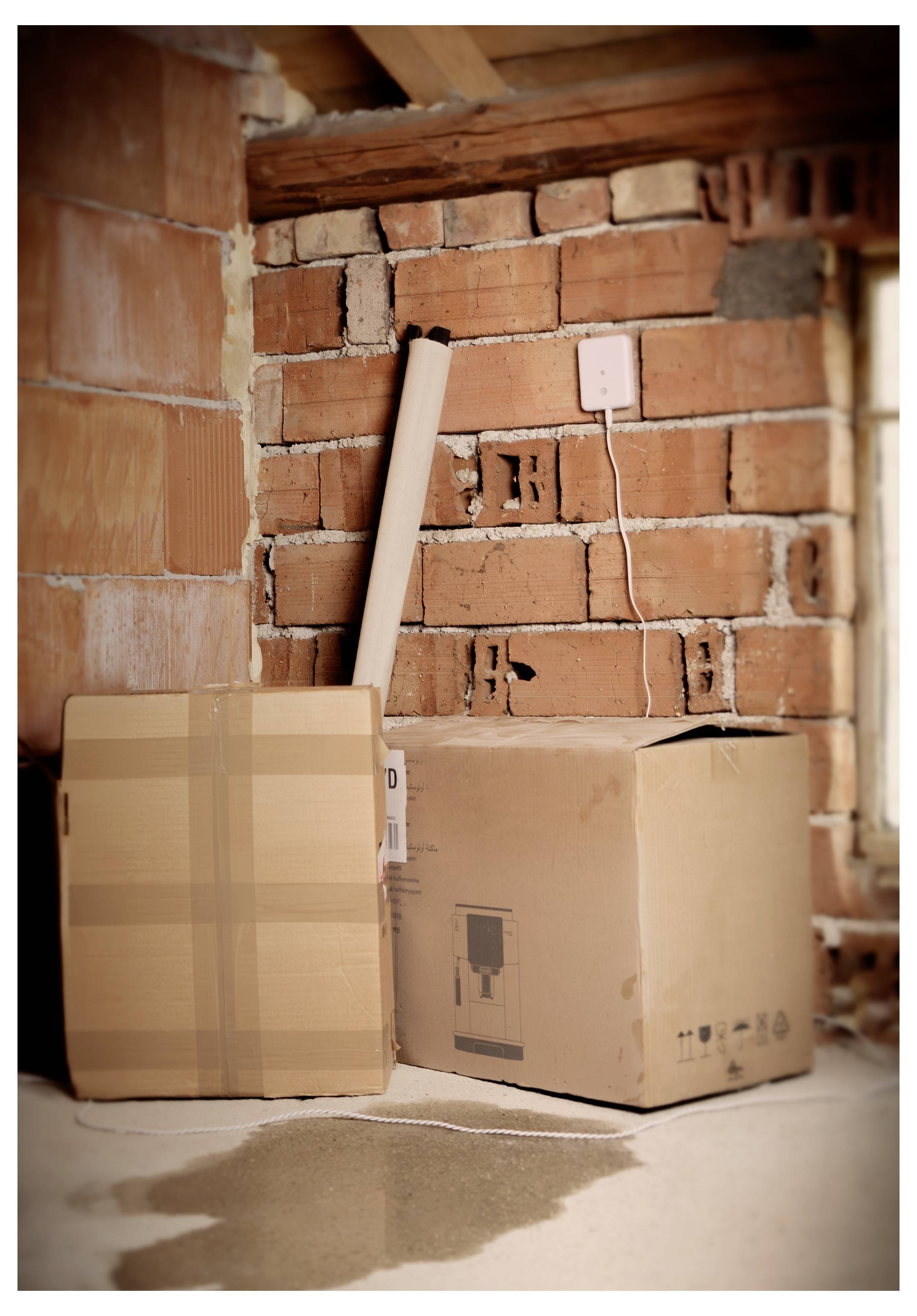 Zwei Pappkartons und ein aufgerollter Teppich in einem teilweise fertiggestellten Raum mit sichtbaren Ziegelwänden und einem Betonboden.