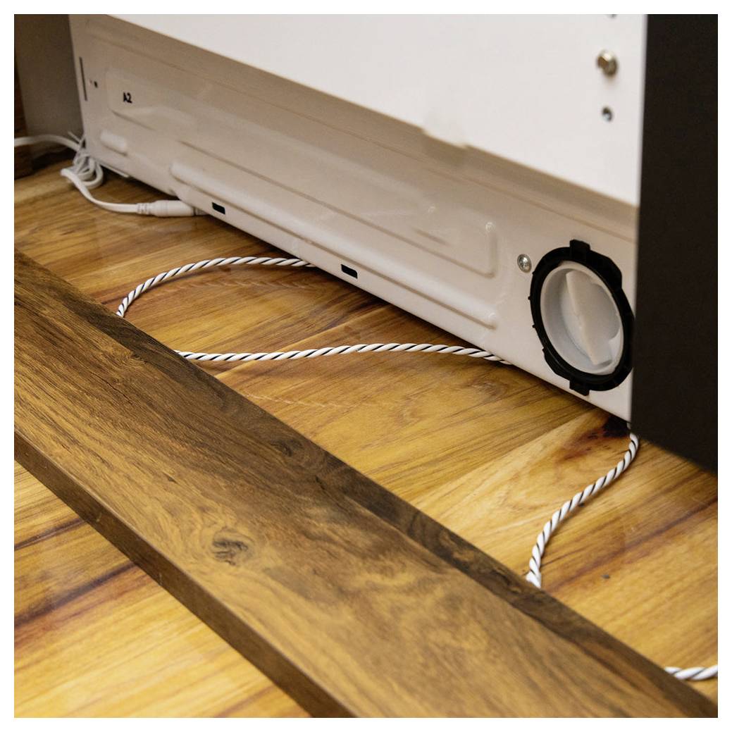 Rand eines Holzbodens, wo er auf eine weiße Geräteabdeckung trifft. Ein Lüftungsschacht mit einem Schlauch ist teilweise sichtbar und deutet auf eine Belüftungsanlage hin.
