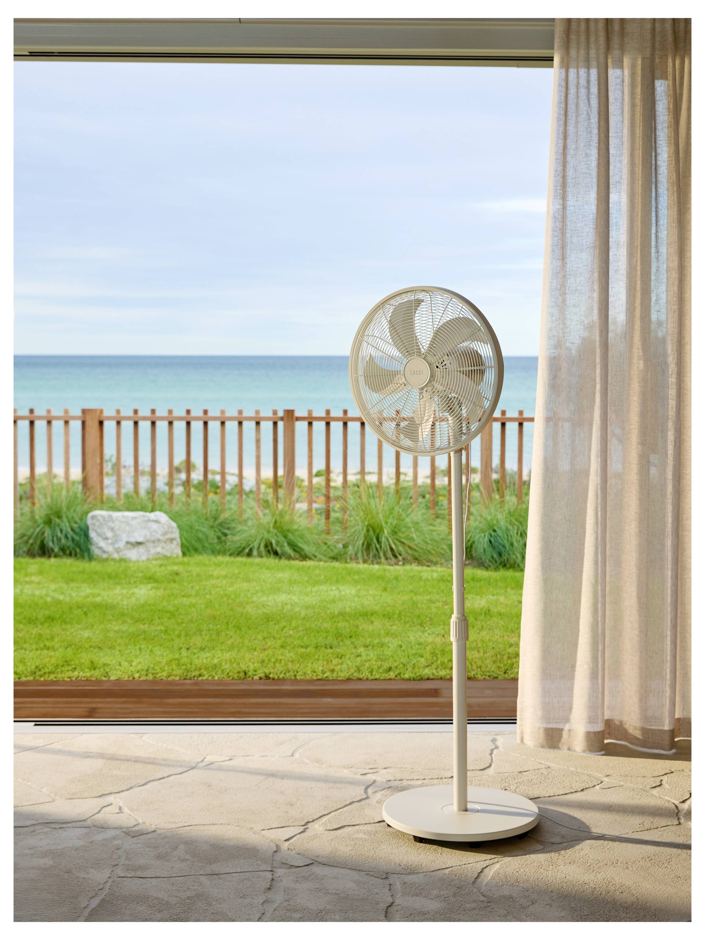 Ein Standventilator in einem Raum blickt auf eine offene Schiebetür, die den Blick auf einen hölzernen Zaun, grünes Gras und ein ruhiges Meer unter einem bewölkten Himmel freigibt.