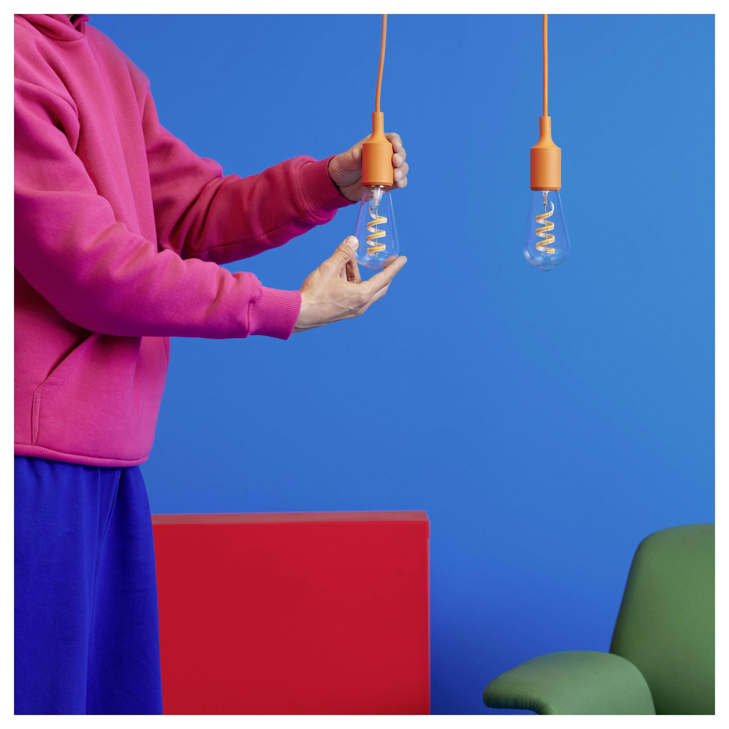 Eine Person in einem rosa Pullover hält zwei hängende orangefarbene Glühbirnen vor einem hellen blau-roten Hintergrund, wobei sich in der Nähe ein grüner Stuhl befindet.