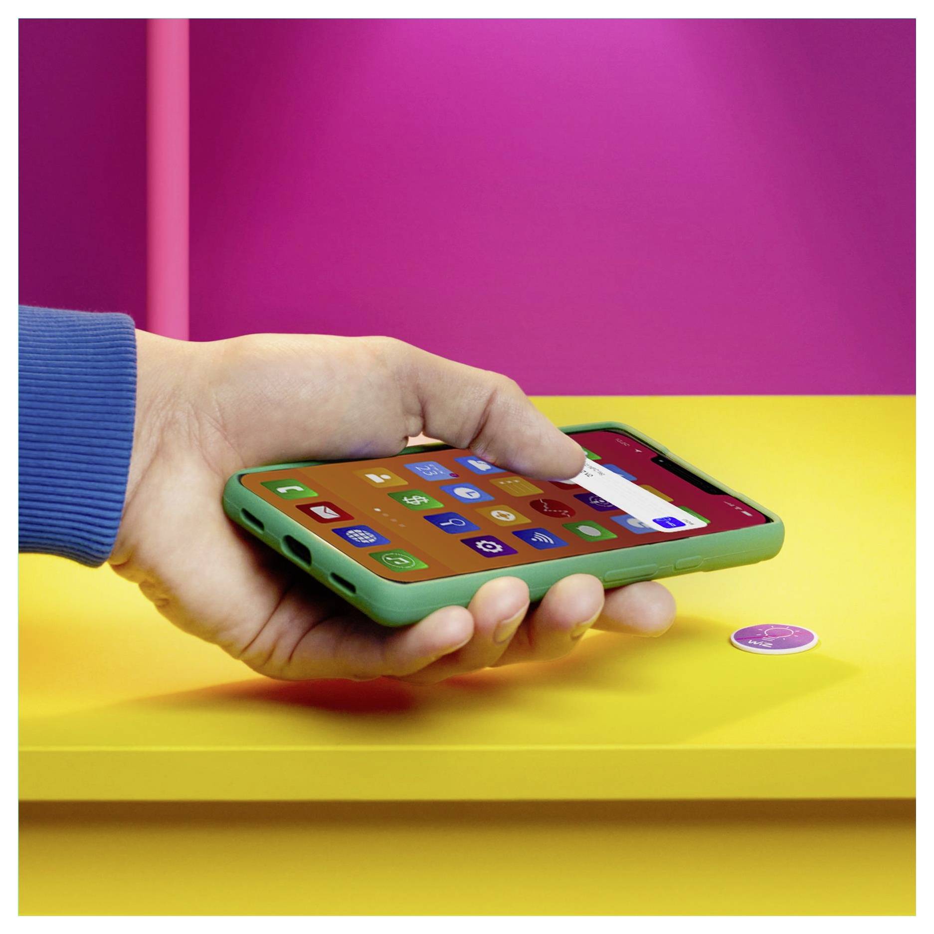 Eine Hand hält ein grünes Smartphone mit bunten App-Symbolen auf dem Bildschirm. Sie schwebt über einem gelben Tisch mit einer pinken Wand im Hintergrund.