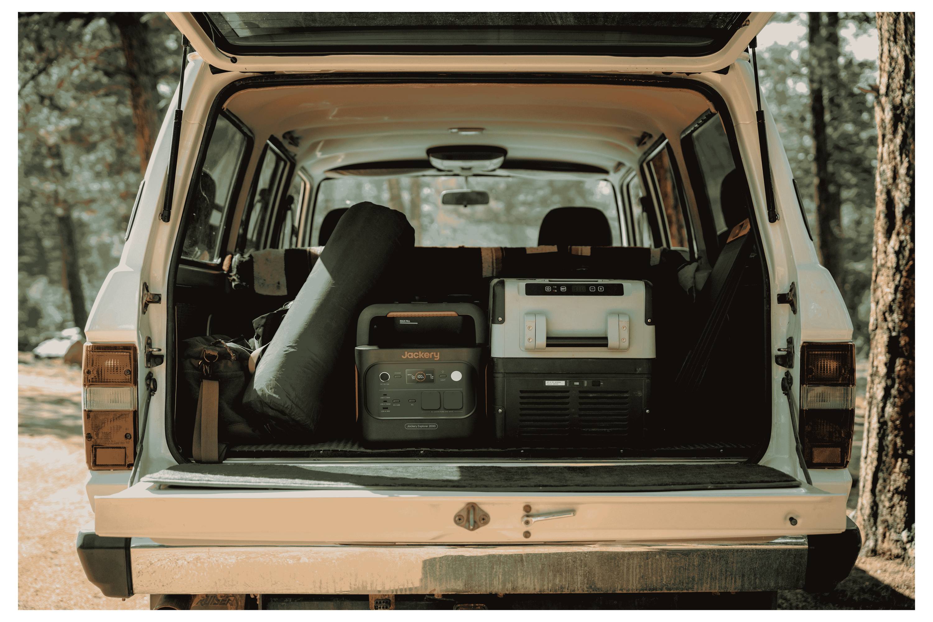 Die Rückseite eines Fahrzeugs ist geöffnet und zeigt Campingausrüstung, darunter einen Kühlschrank, eine Powerstation und einen zusammengerollten Schlafsack, vor einer Waldkulisse.