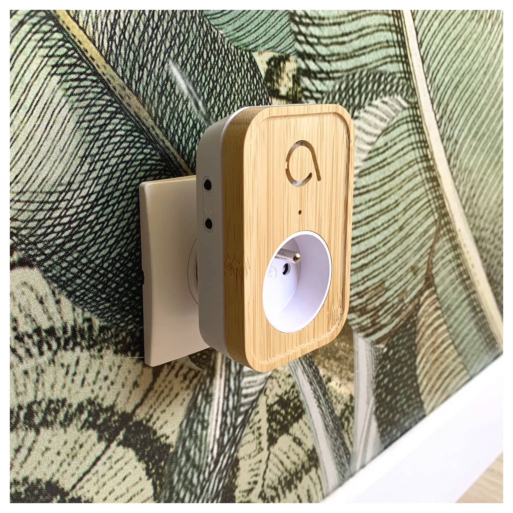 Eine intelligente Steckdose mit Bambusoptik ist in eine Wandsteckdose eingesteckt, vor dem Hintergrund einer grün-weiß gemusterten Tapete.