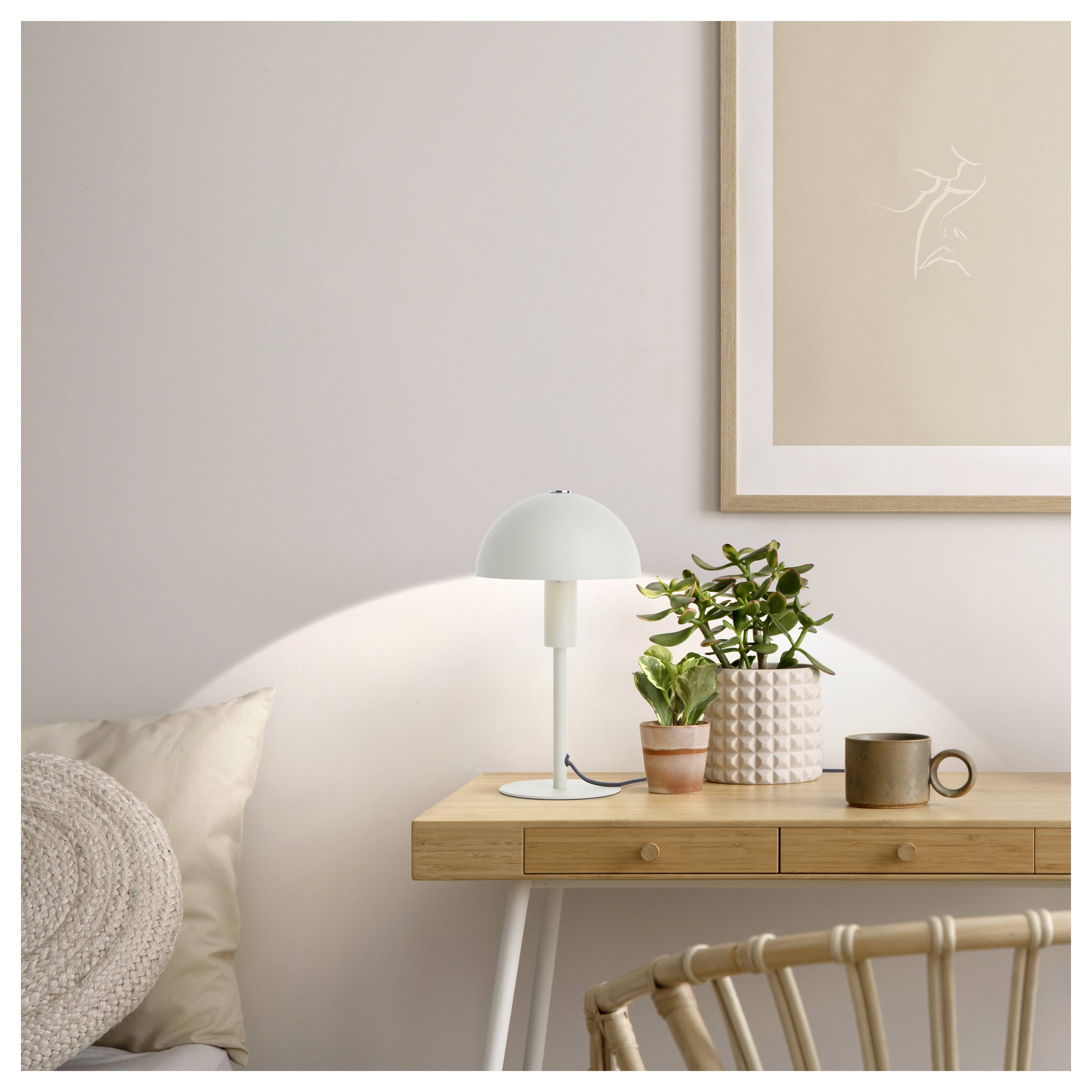Ein minimalistischer Holzschreibtisch mit einer Zimmerpflanze, einer weißen Kuppellampe und einem braunen Becher, vor einer neutralen Wand mit abstrakter Kunst.