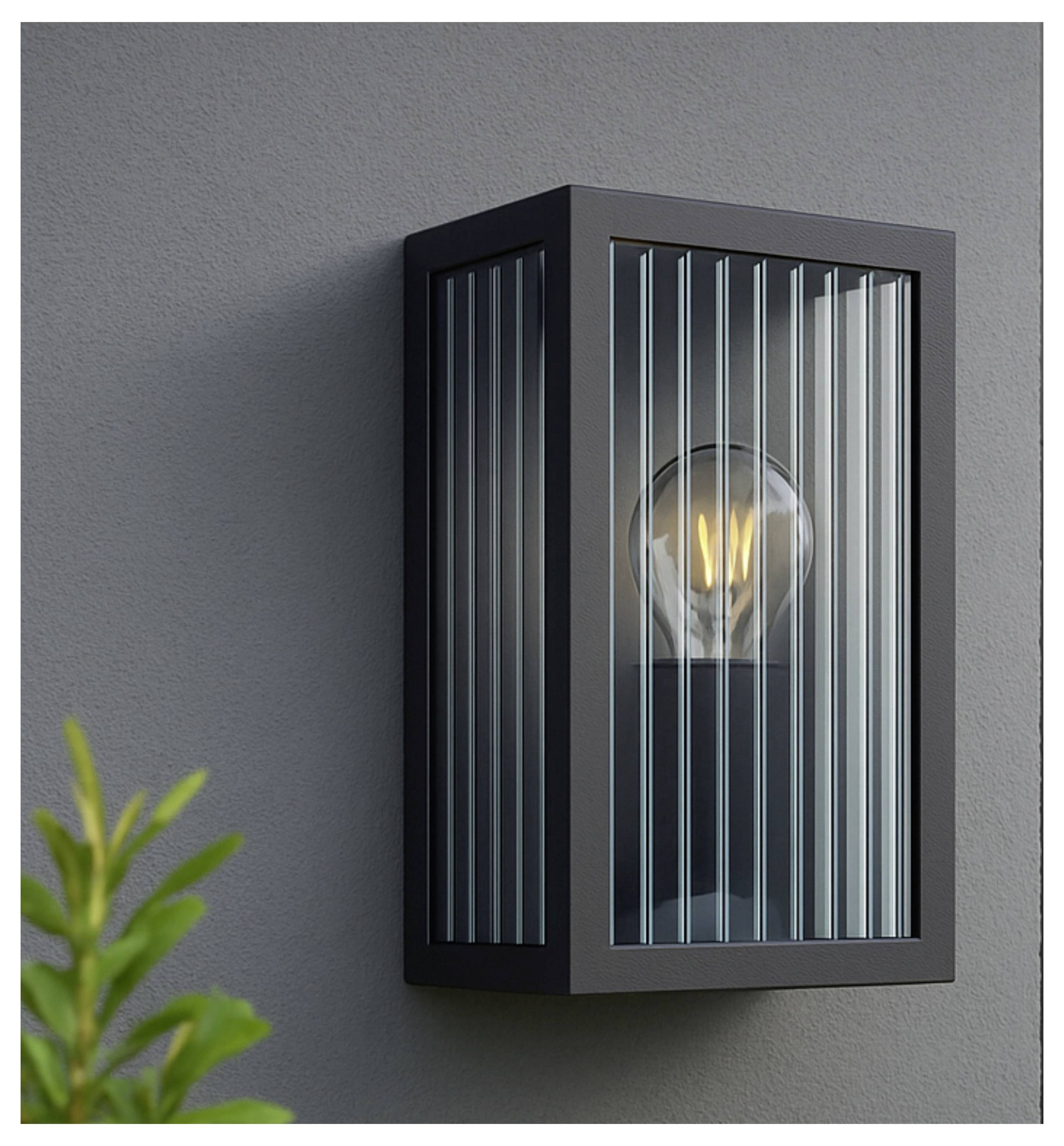 Eine moderne Außenwandlampe mit einer in Glas eingeschlossenen Glühbirne, die an einer grauen Wand montiert ist, neben einigen grünen Blättern auf der linken Seite.