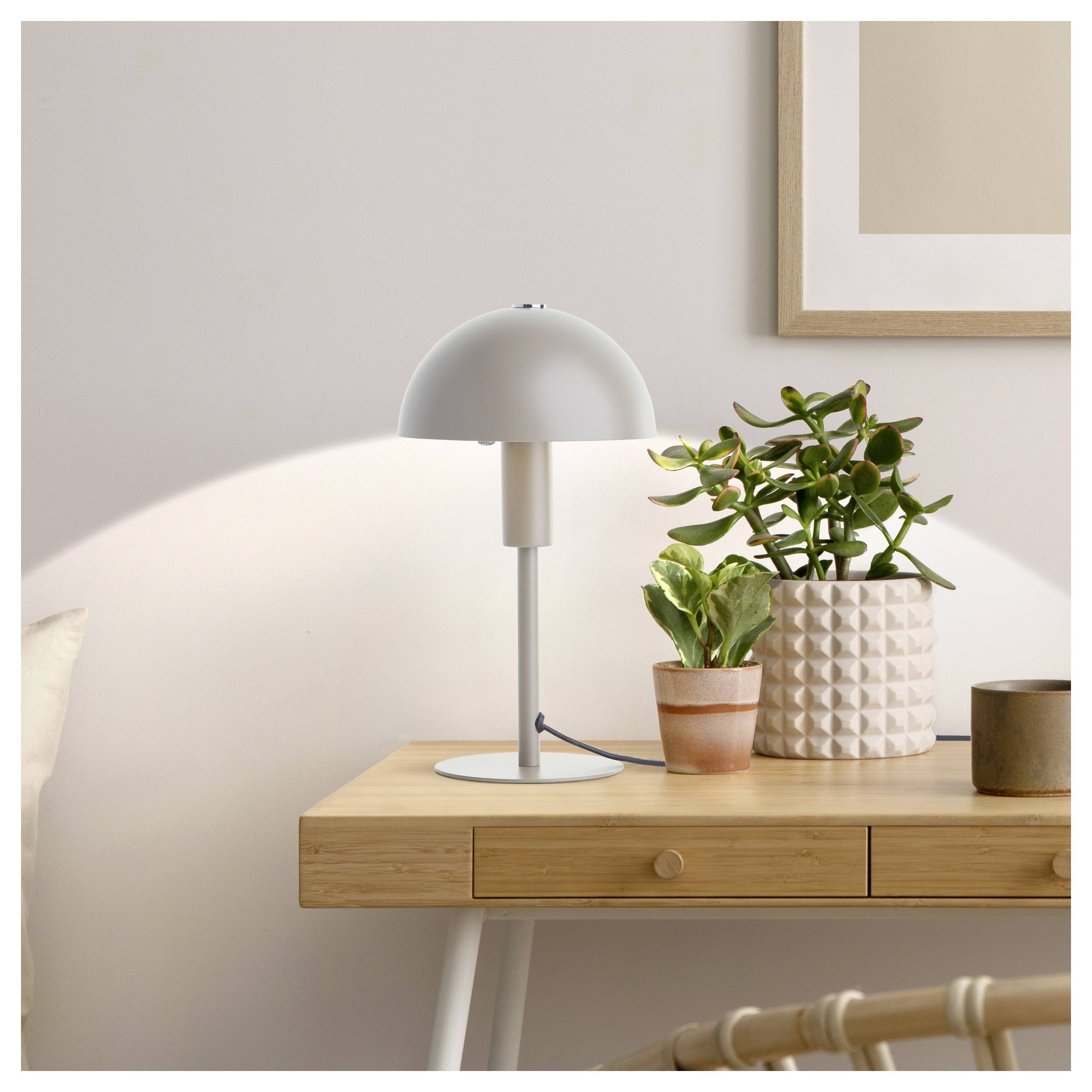 Ein minimalistisches Zimmer mit einer angeschalteten weißen Schreibtischlampe, einer Zimmerpflanze und einem kleinen Holzbehälter auf einem Holztisch vor einer hellen Wand.