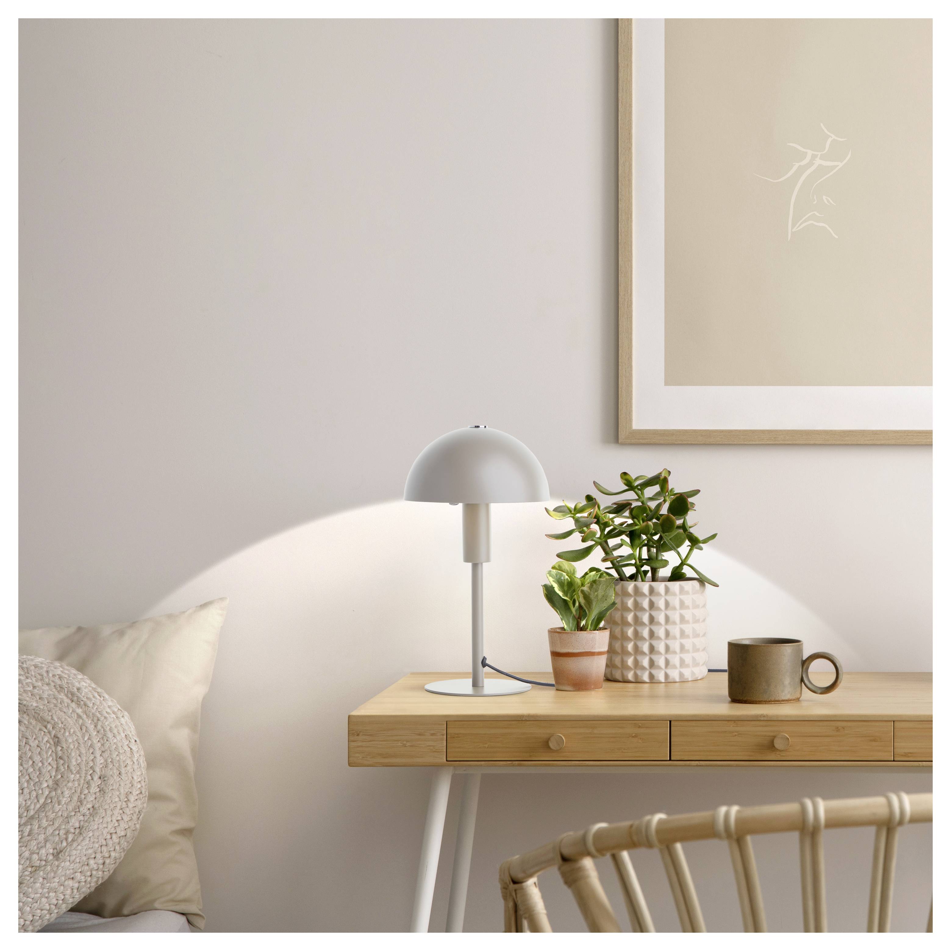 Holzschreibtisch mit einer kleinen Lampe, einer Topfpflanze und einer Tasse. Beige Wandkunst und ein Kissen auf dem Stuhl tragen zu der gemütlichen minimalistischen Atmosphäre bei.