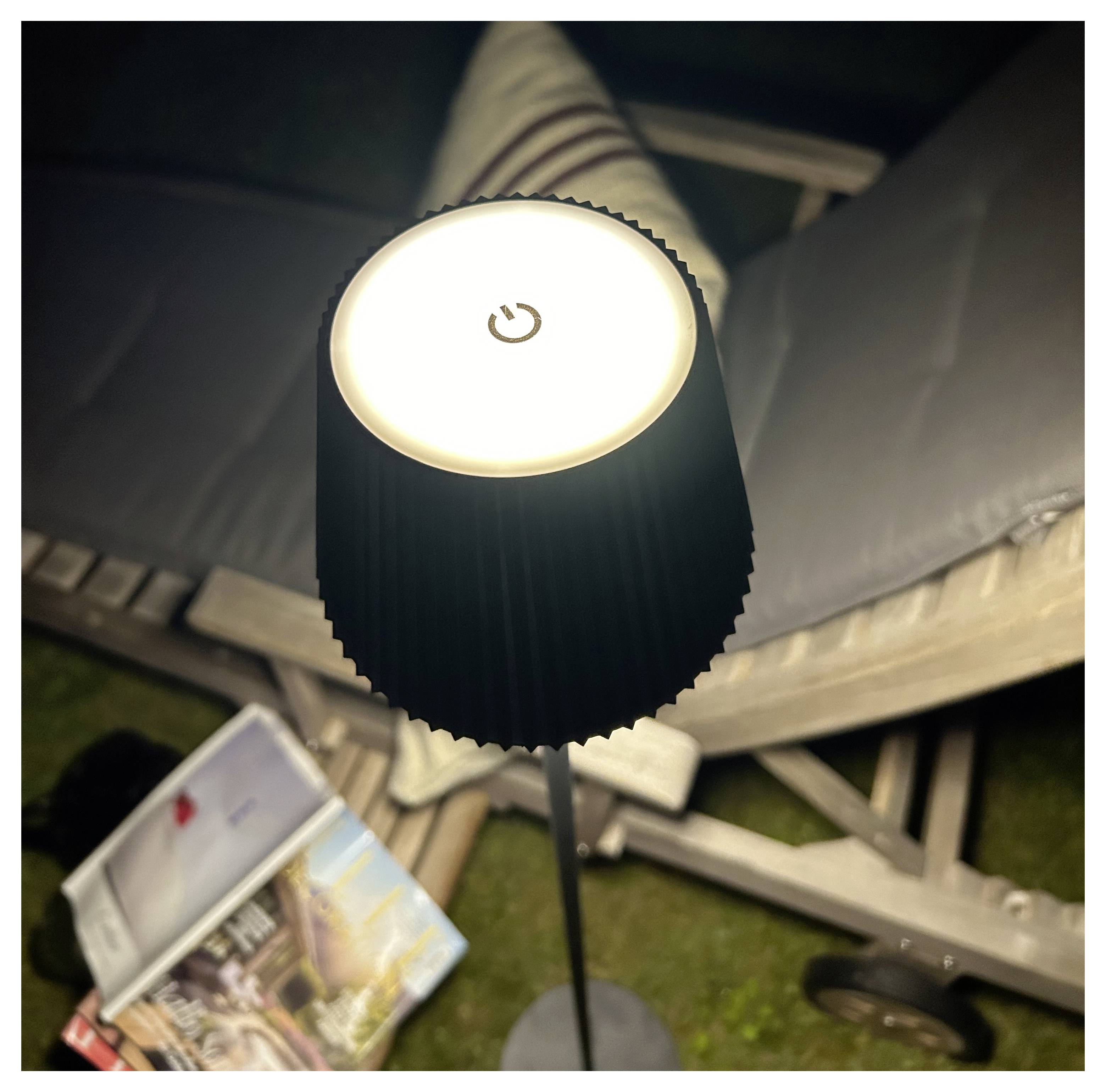 Eine stehende LED-Lampe mit einer Netzschalter-Taste auf der Oberseite, die einen Bereich mit Zeitschriften auf einem Stuhl und einem Kissen ausleuchtet und einen gemütlichen Lesebereich andeutet.