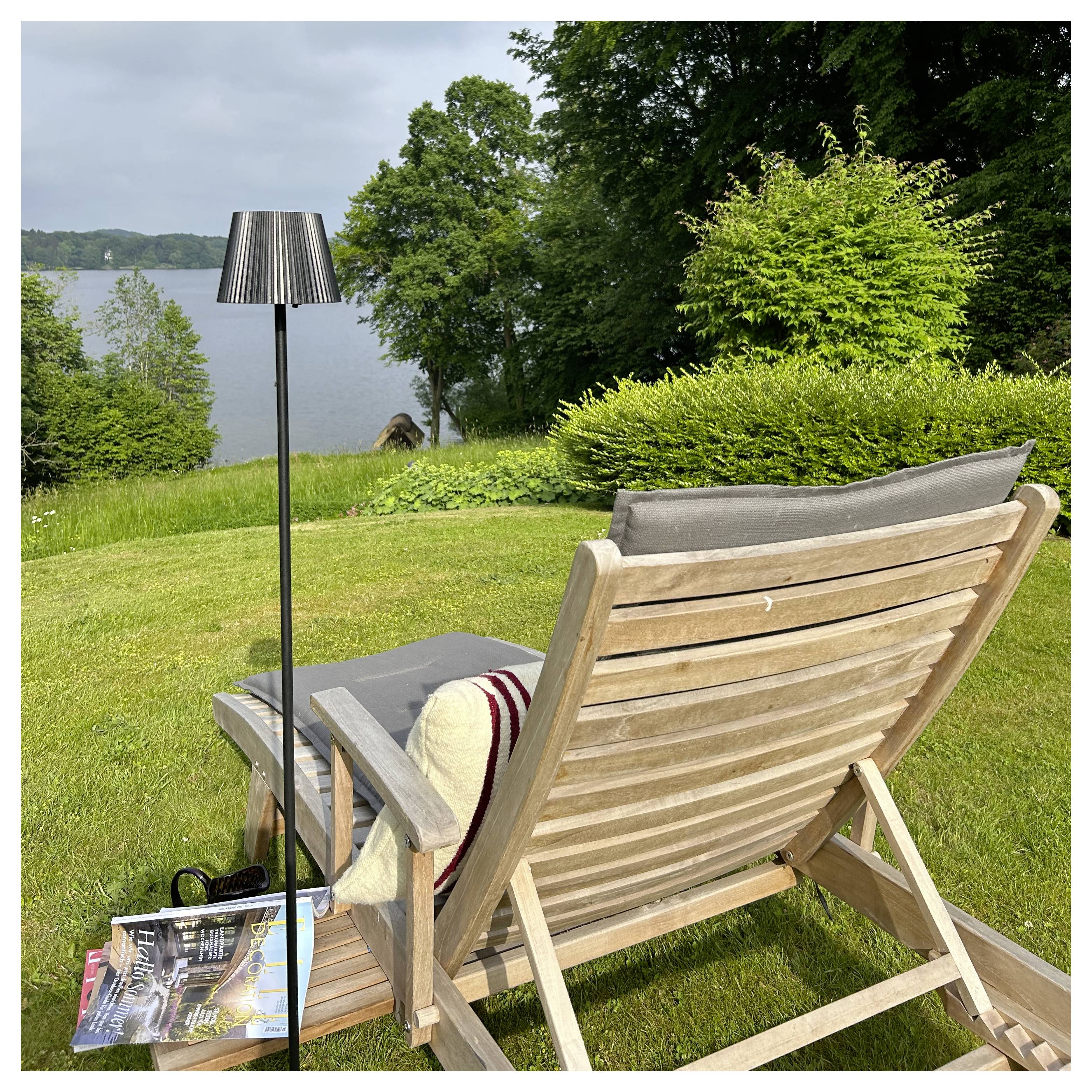 Ein Liegestuhl aus Holz mit einem Kissen und einer Decke blickt auf einen Seeblick, umgeben von üppiger Vegetation und einer Lampe an seiner Seite.