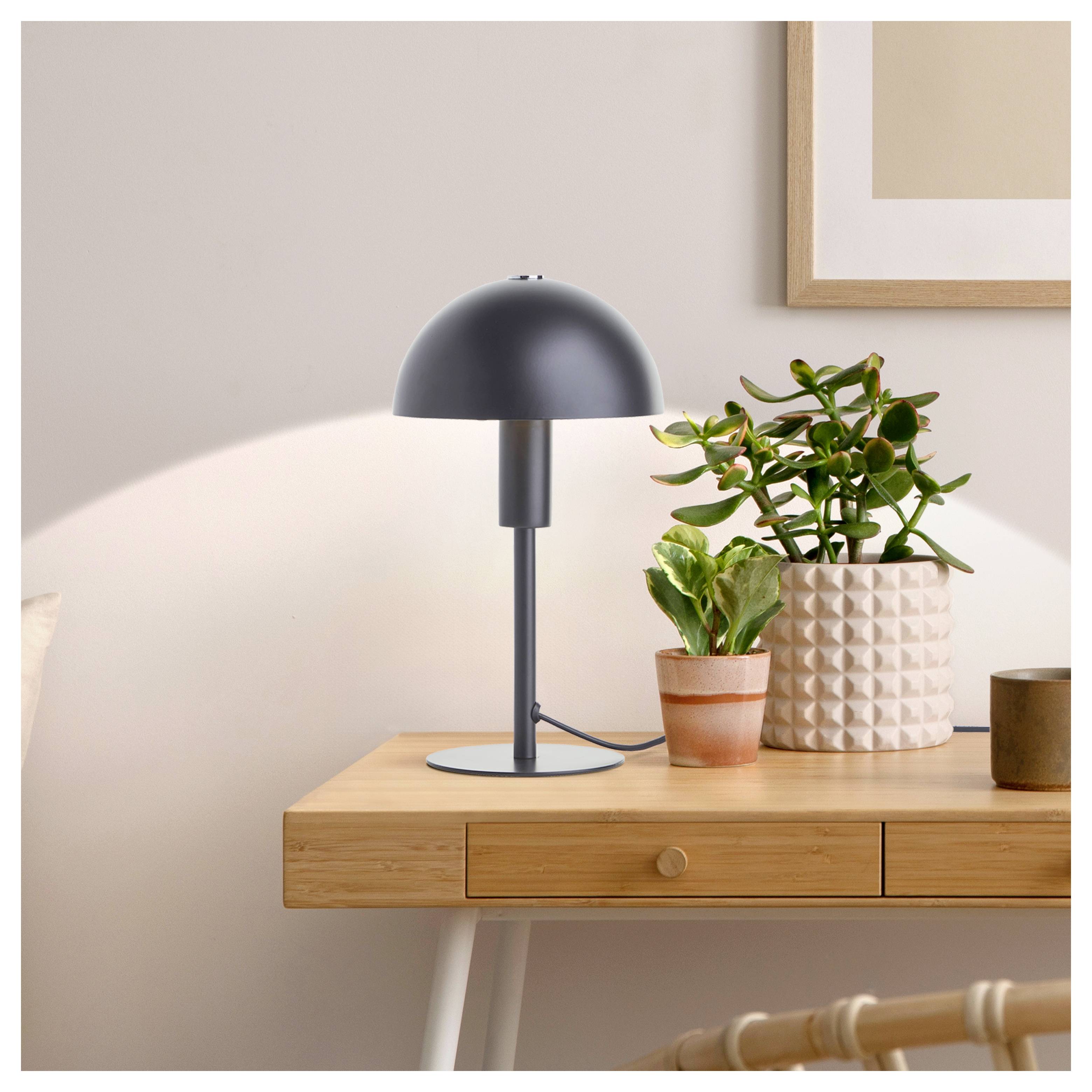 Eine elegante schwarze Tischlampe auf einem Holzschreibtisch neben einer Topfpflanze, die ein warmes Licht in einem modern-minimalistischen Raum verbreitet.