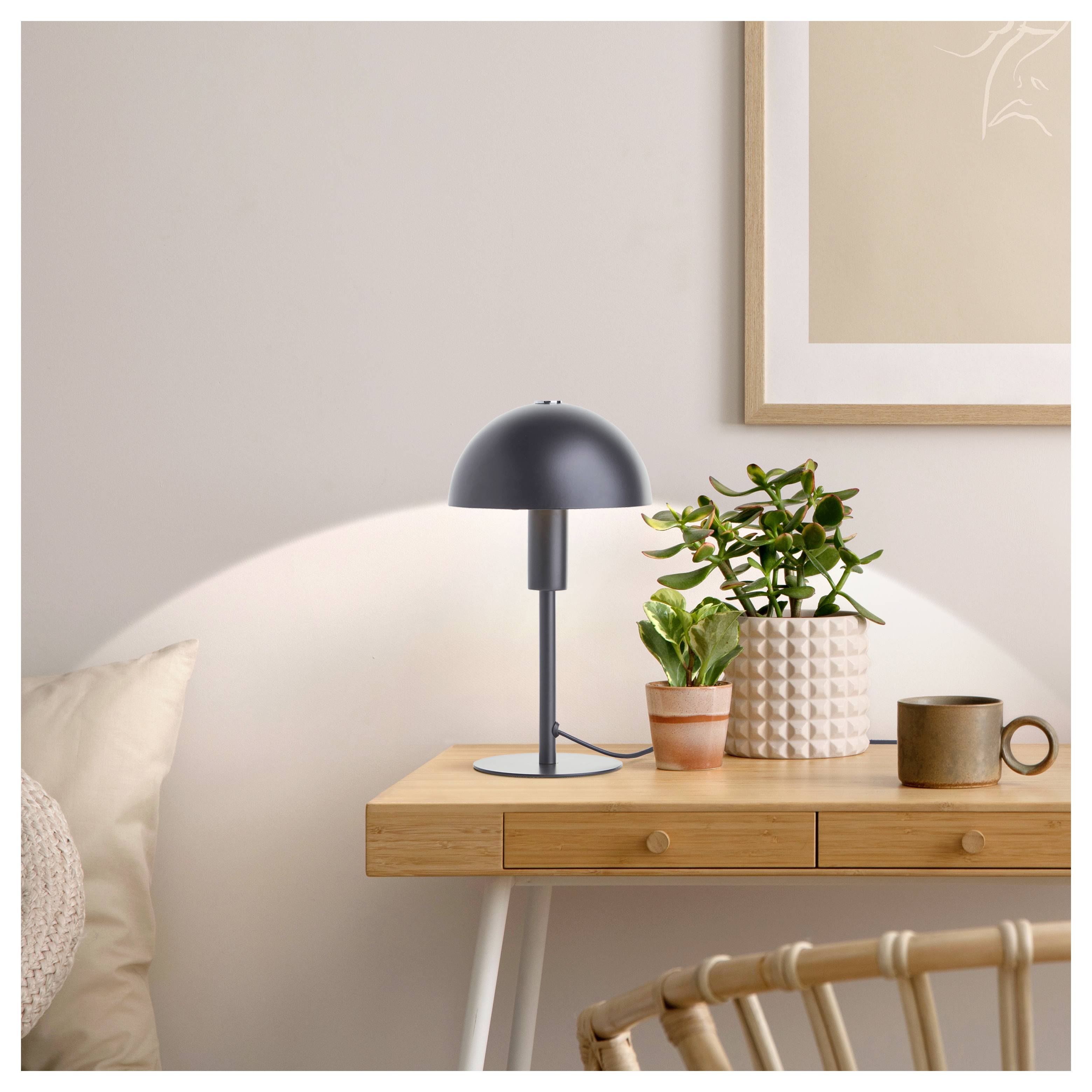 Ein Holztisch trägt eine moderne Lampe, eine Topfpflanze und einen Becher; darüber hängt ein gerahmtes Bild. Gemütliche, minimalistische Raumgestaltung.