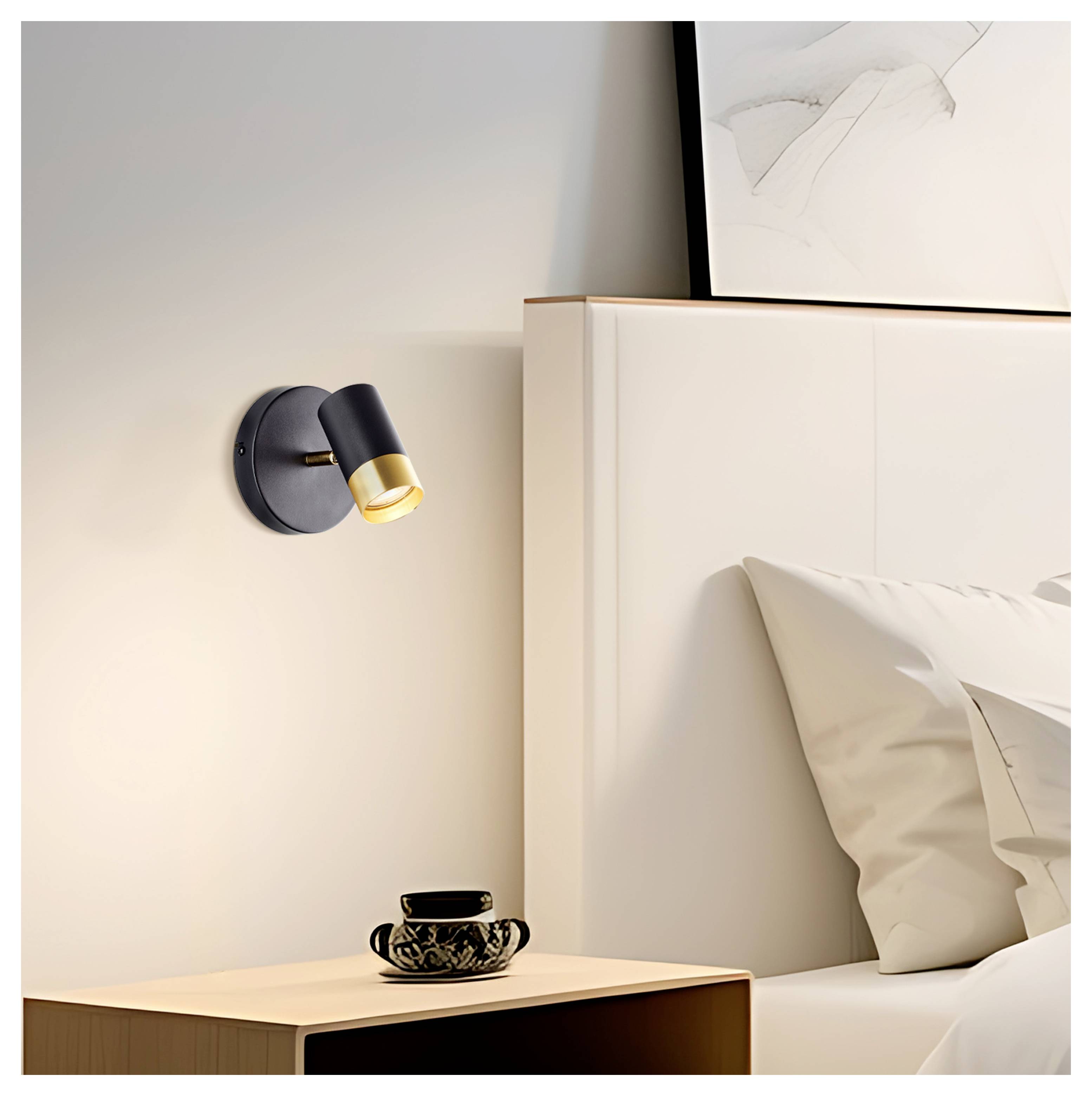 Eine moderne Wandlampe beleuchtet einen Schlafbereichs mit einem Holznachttisch, auf dem eine schwarze Tasse steht, neben einem weißen Bett und abstrakter Kunst.