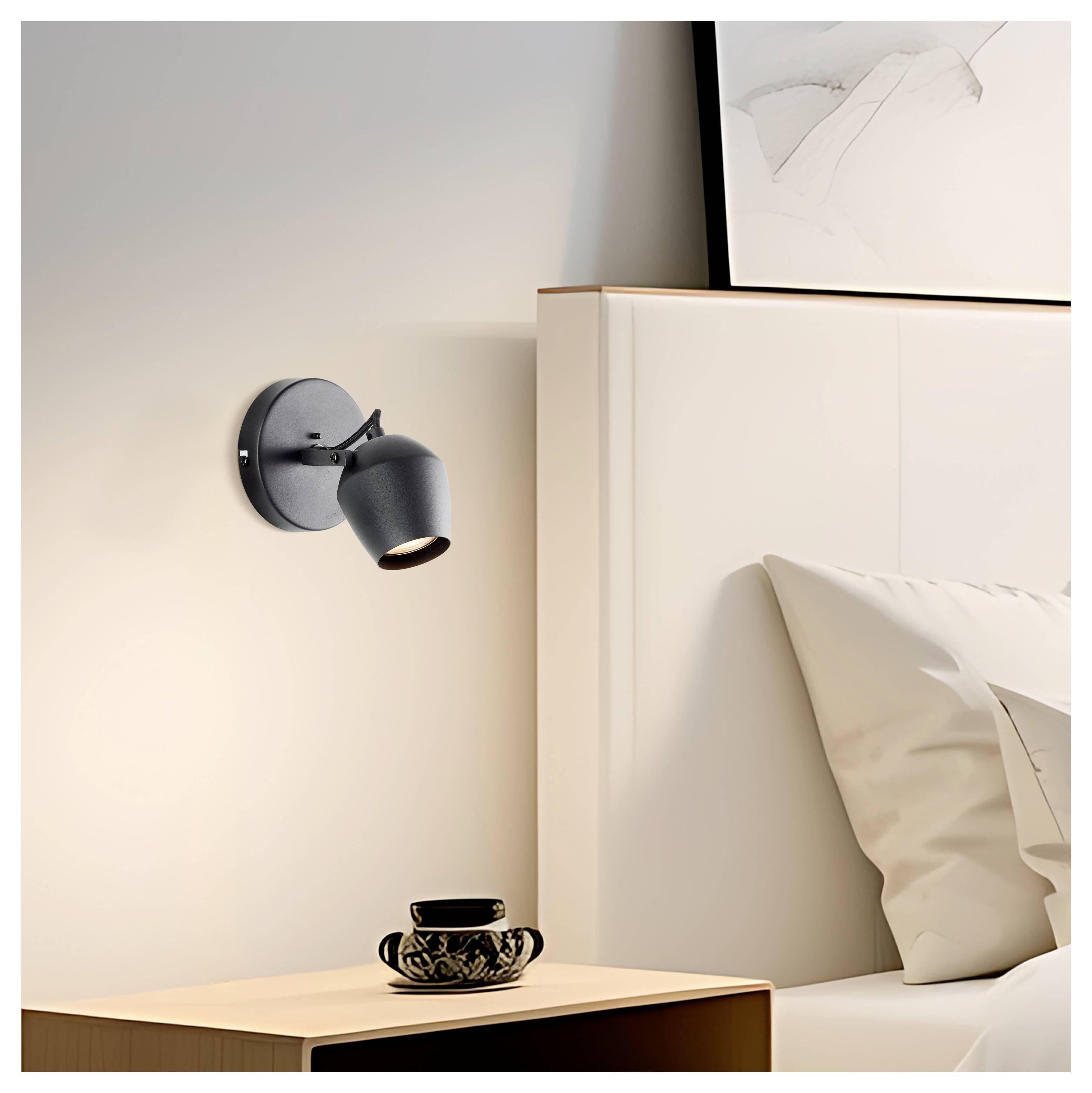 Ein modernes Schlafzimmer mit einer wandmontierten schwarzen Leselampe über einem Holznachttisch. Eine elegante Teetasse steht auf dem Nachttisch; über dem Bett ist ein gerahmtes Kunstwerk zu sehen.