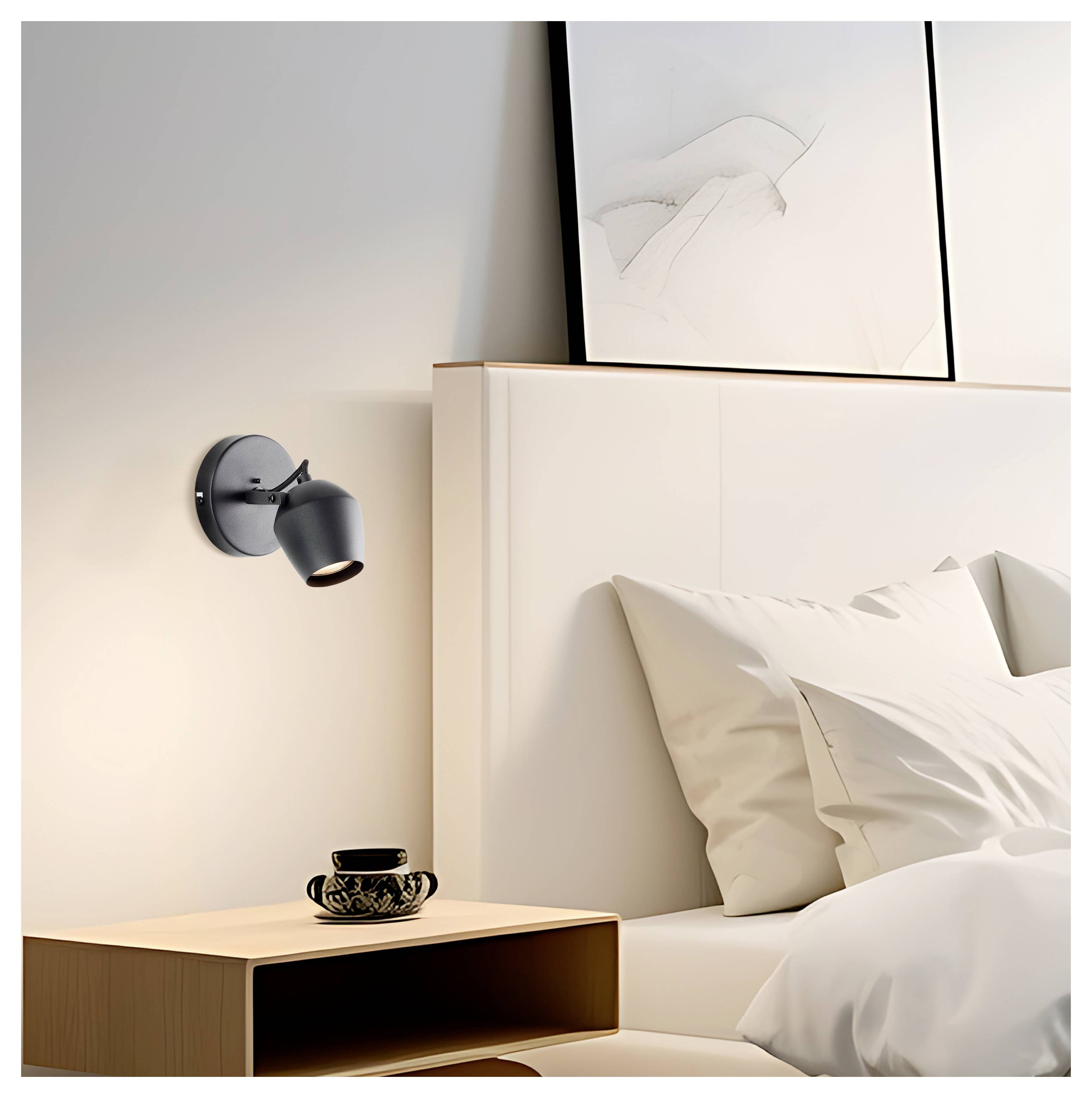 Eine gemütliche Ecke eines Schlafzimmers mit einem Scheinwerfer, der auf eine dekorative Teetasse auf einem Holznachttisch scheint, und einem minimalistischen gerahmten Kunstwerk über dem Bett.