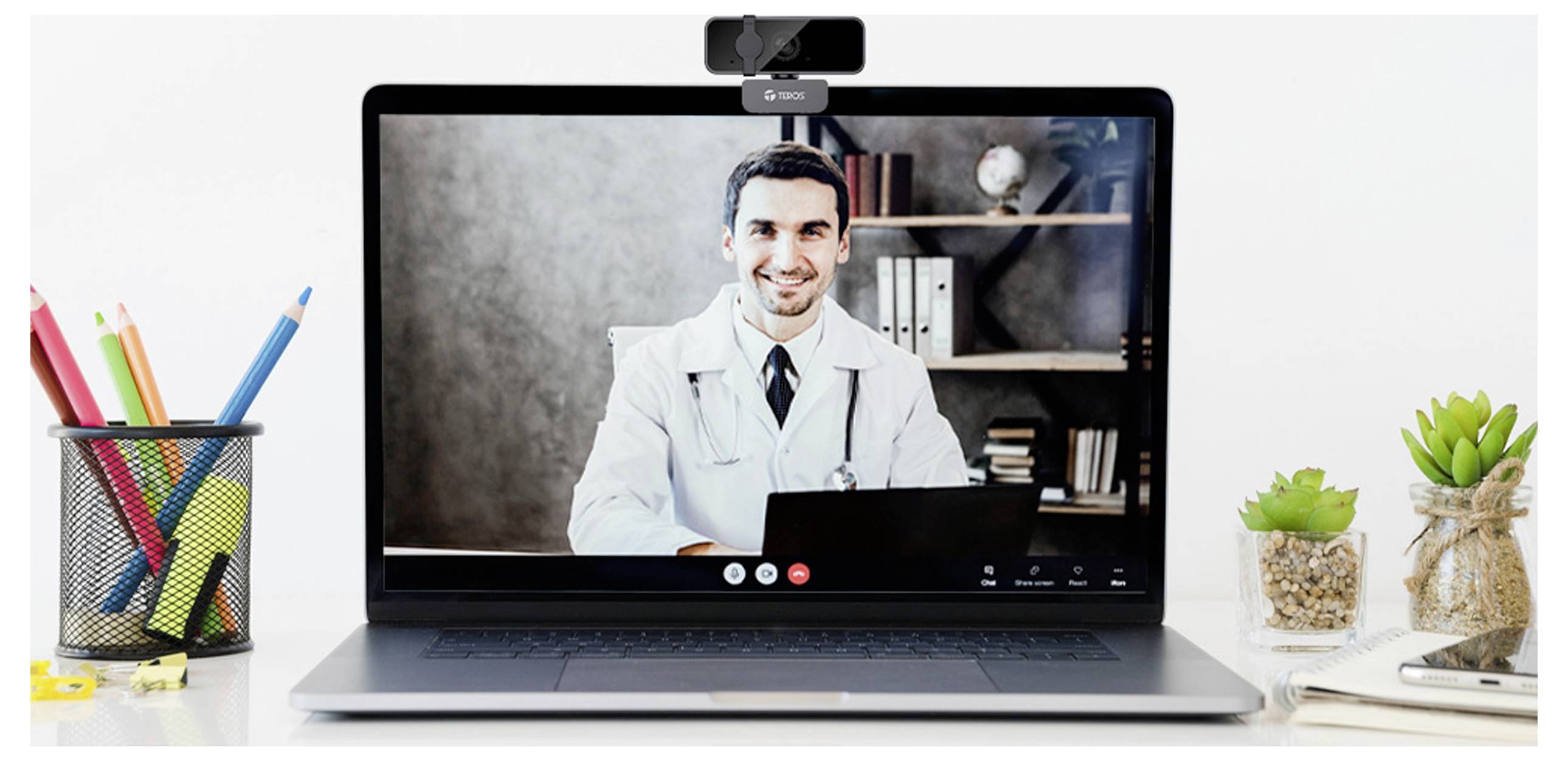 Ein Arzt in einem weißen Kittel führt eine Videokonferenz auf einem Laptop, umgeben von Büroartikeln, darunter Pflanzen und ein Stifthalter.