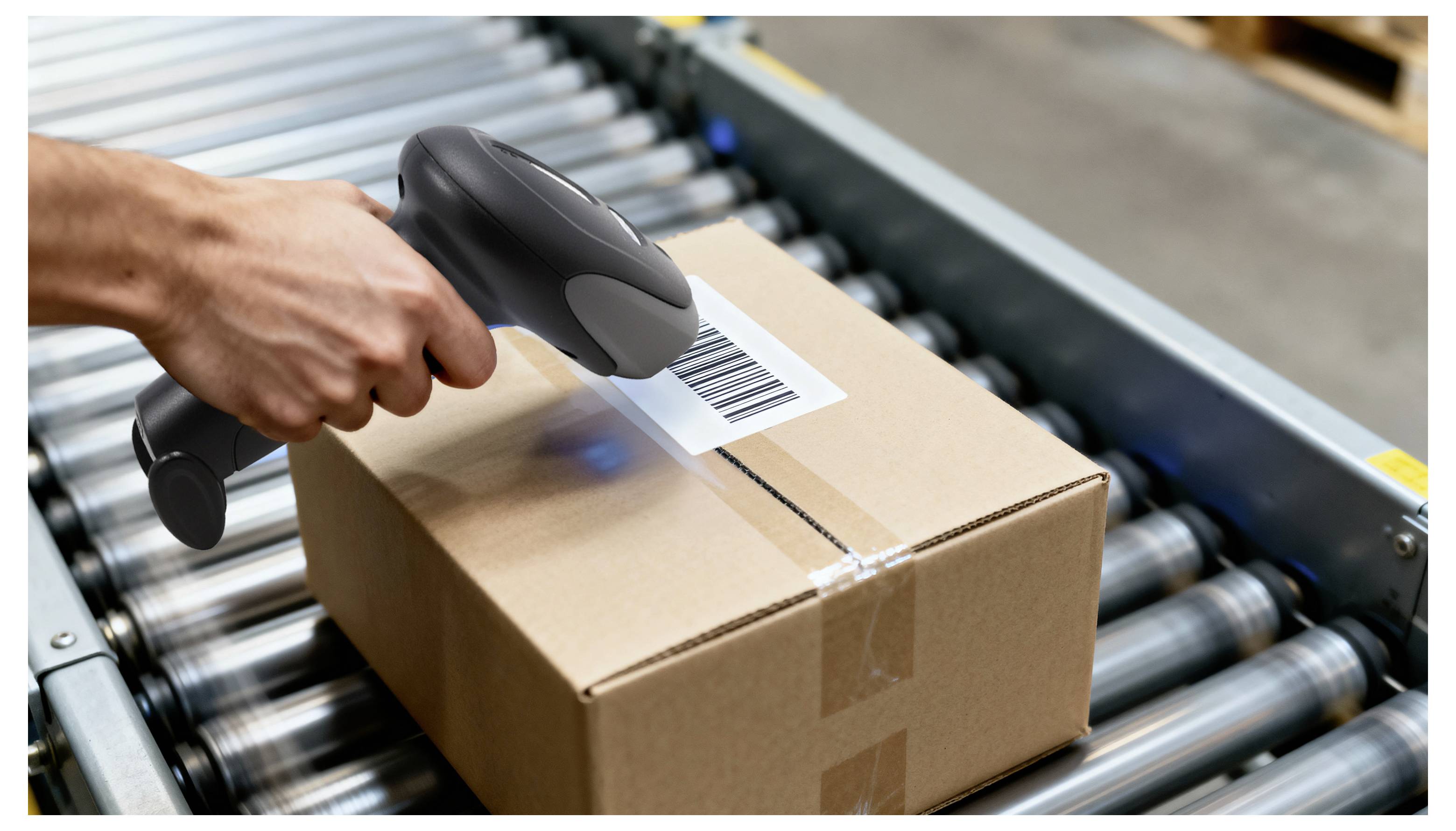 Eine Hand scannt einen Barcode auf einem Karton mit einem tragbaren Scanner. Der Karton befindet sich auf einem Förderband in einem Lagerhaus.