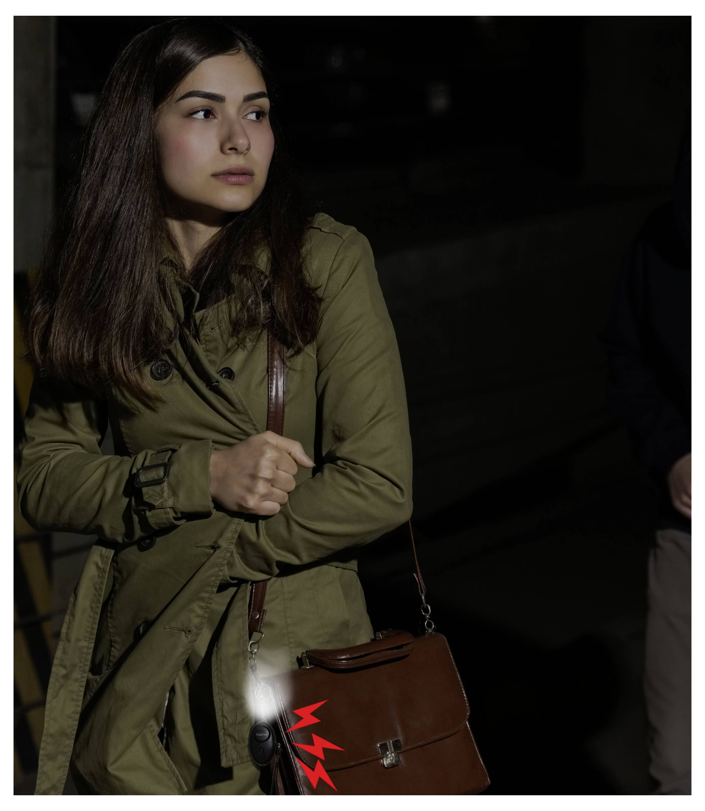 Frau in einem olivfarbenen Trenchcoat, die eine braune Umhängetasche hält, steht in schwachem Licht und blickt zur Seite.