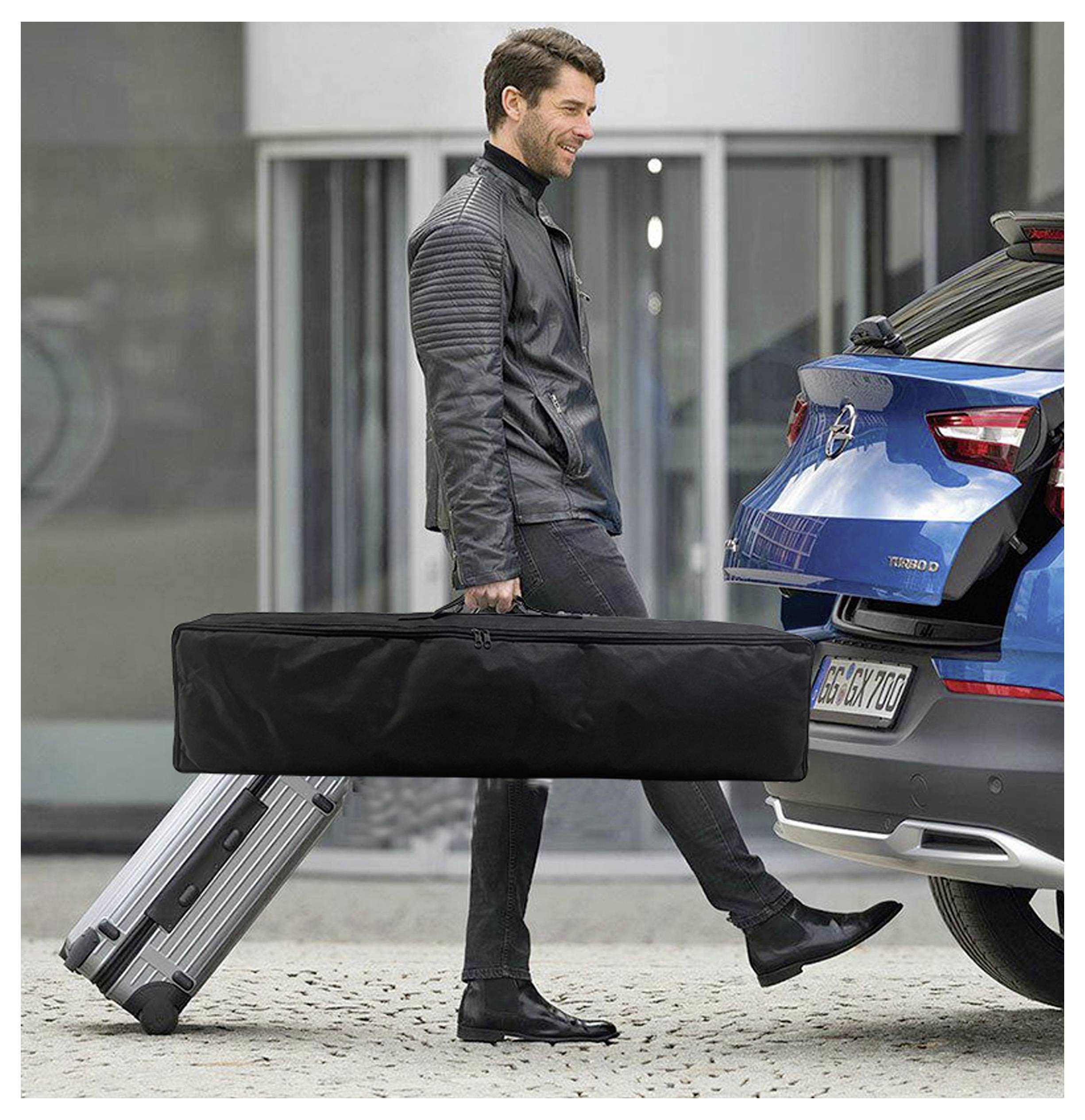 Mann in einer schwarzen Jacke, der einen großen Koffer zum Heck eines blauen Autos fährt, als würde er Gepäck laden oder entladen.