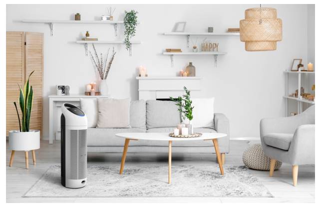 Ein gemütliches Wohnzimmer mit einem grauen Sofa, einem weißen Couchtisch und Topfpflanzen. Ein moderner Luftreiniger ist prominent im Vordergrund platziert.