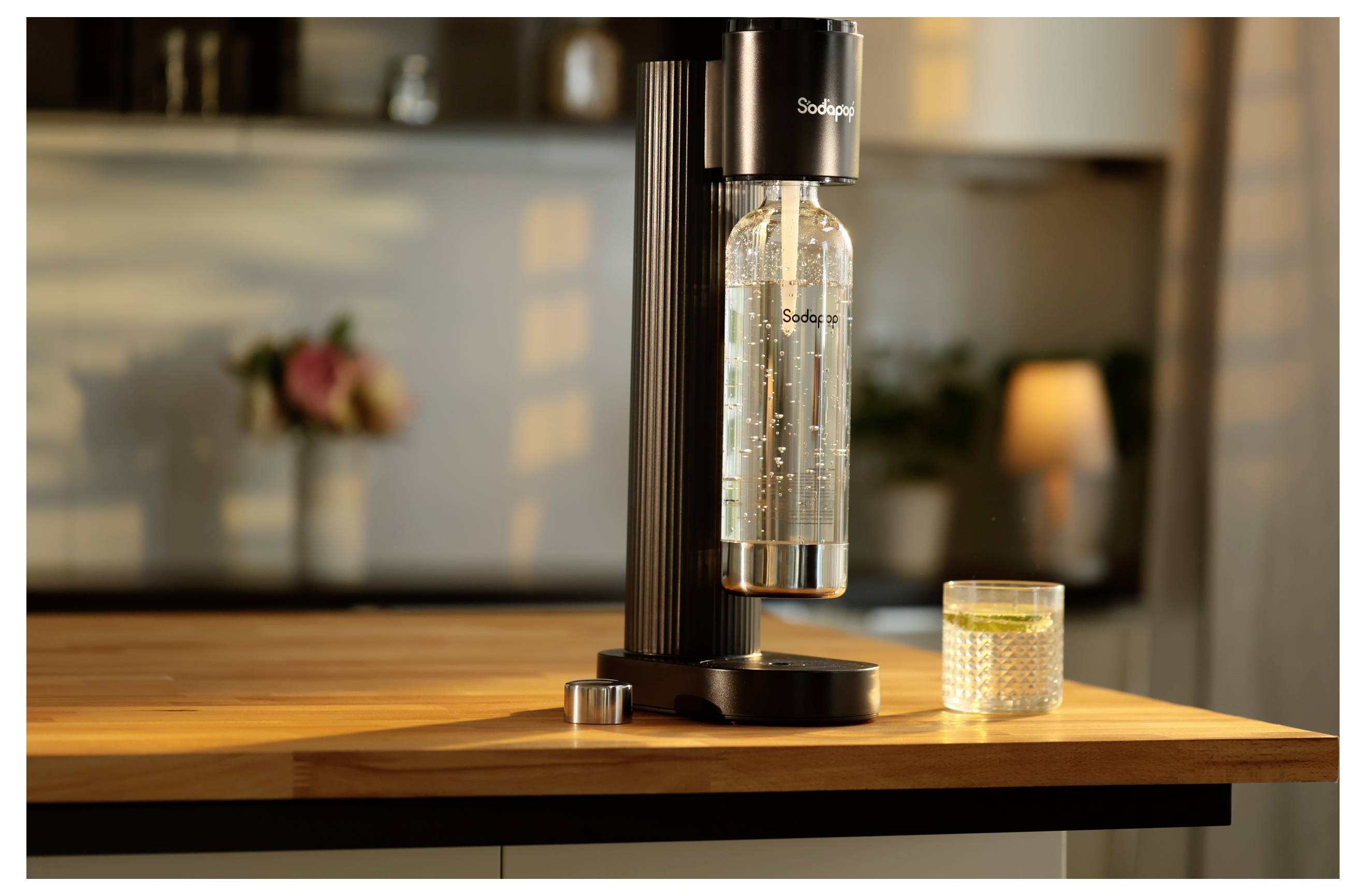 Ein schwarzer Sodahersteller auf einer Küchenarbeitsplatte mit einer transparenten Flasche angeschlossen, neben einem gefüllten texturierten Glas, in einer warm beleuchteten Küchenumgebung.