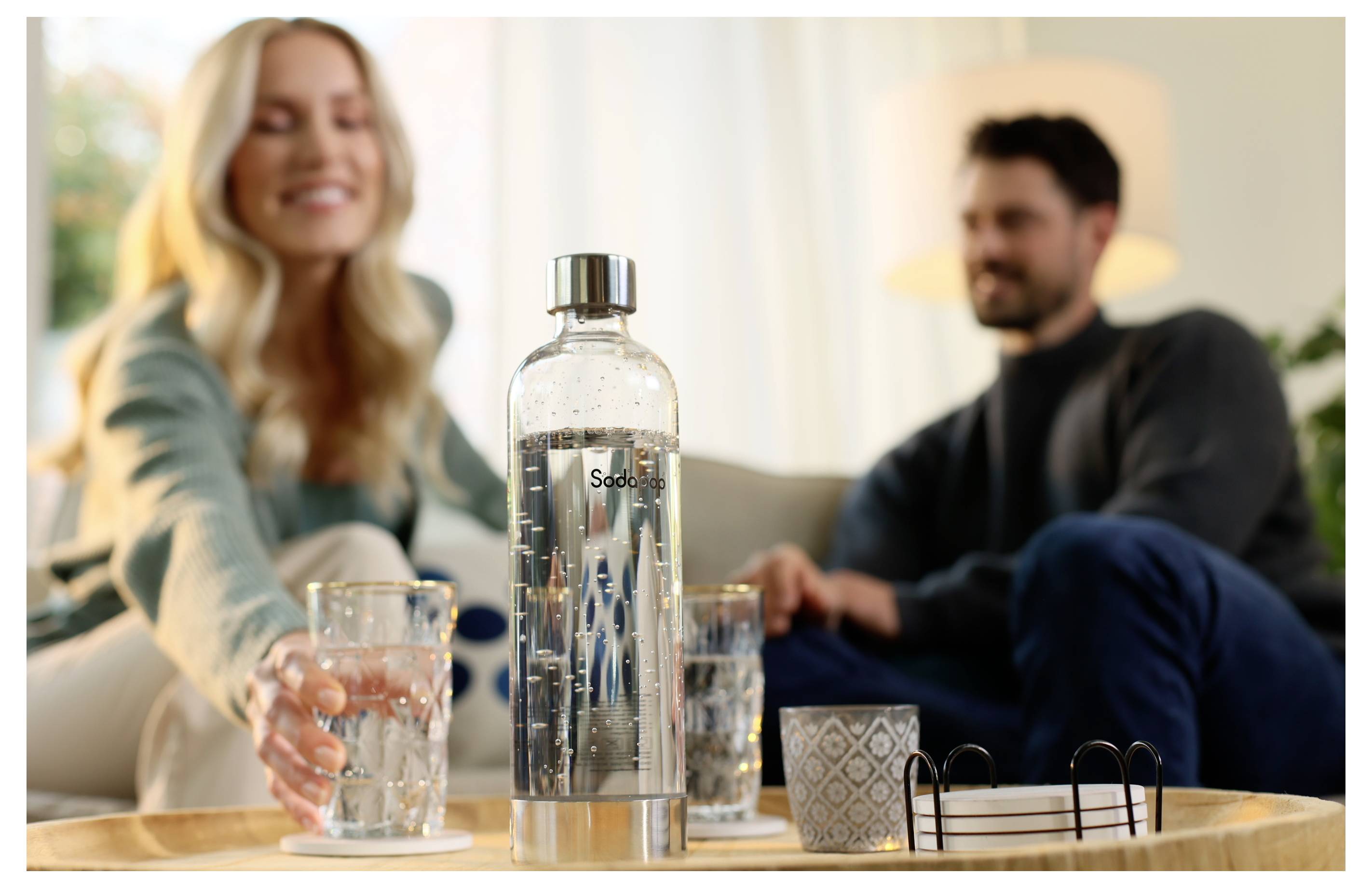 Eine Frau, die einem Mann ein Glas Sprudelwasser auf einem Tablett mit einer SodaStream-Flasche und dekorativen Elementen in einem gemütlichen Wohnzimmer reicht.