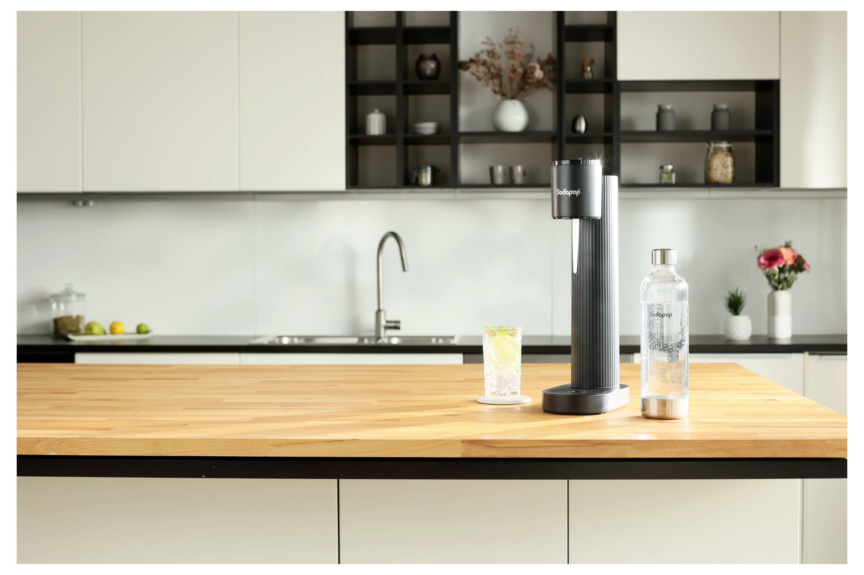Eine moderne Küchenarbeitsplatte mit einem Sprudelwasserbereiter, einem Glas Zitronenwasser und einer Flasche. Im Hintergrund Regale mit Dekoartikeln.