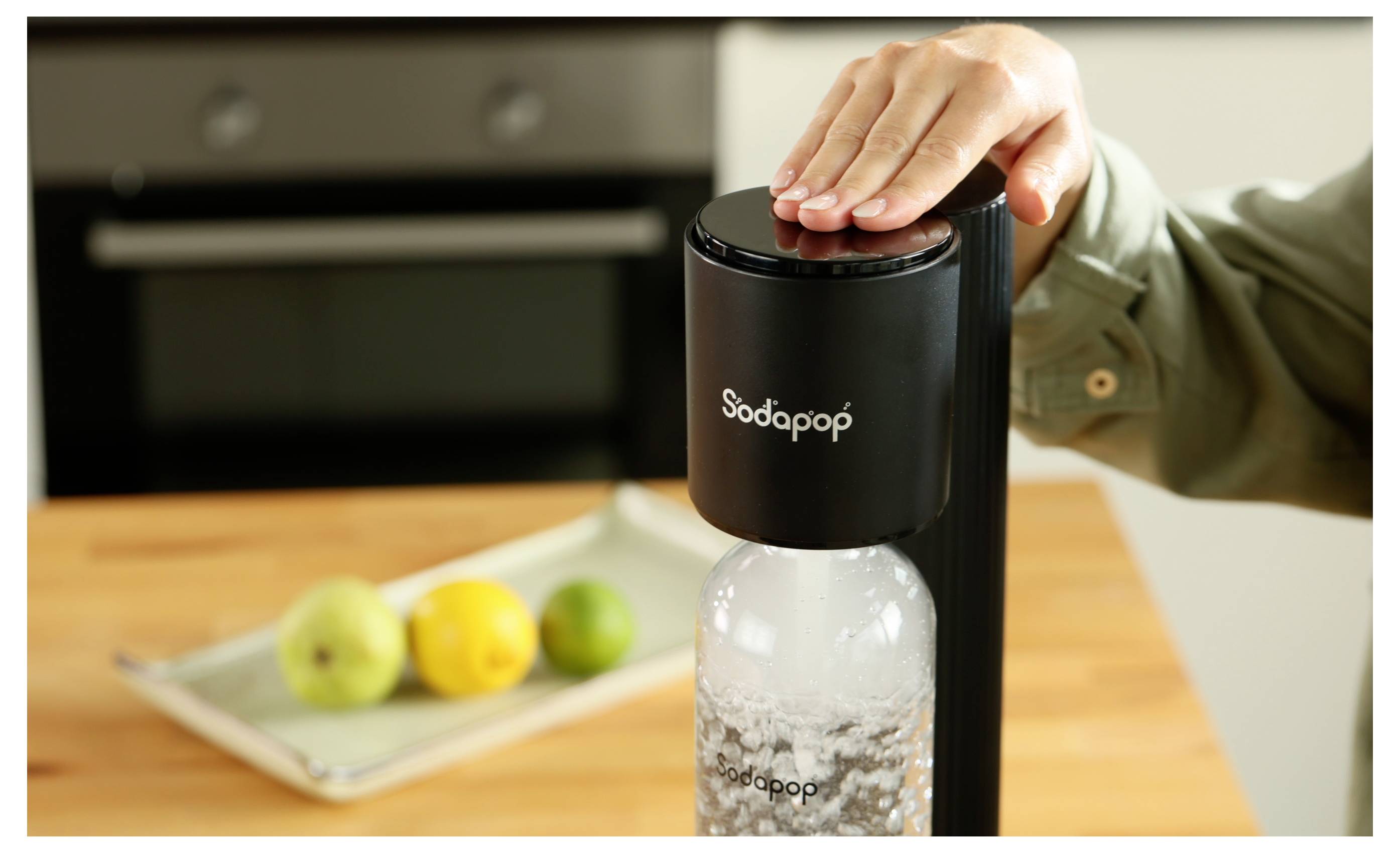 Eine Hand drückt auf den Deckel einer Sodapop-Maschine und karbonisiert Wasser in einer Flasche. Ein Tablett mit Obst ist im Hintergrund auf der Küchenarbeitsplatte zu sehen.