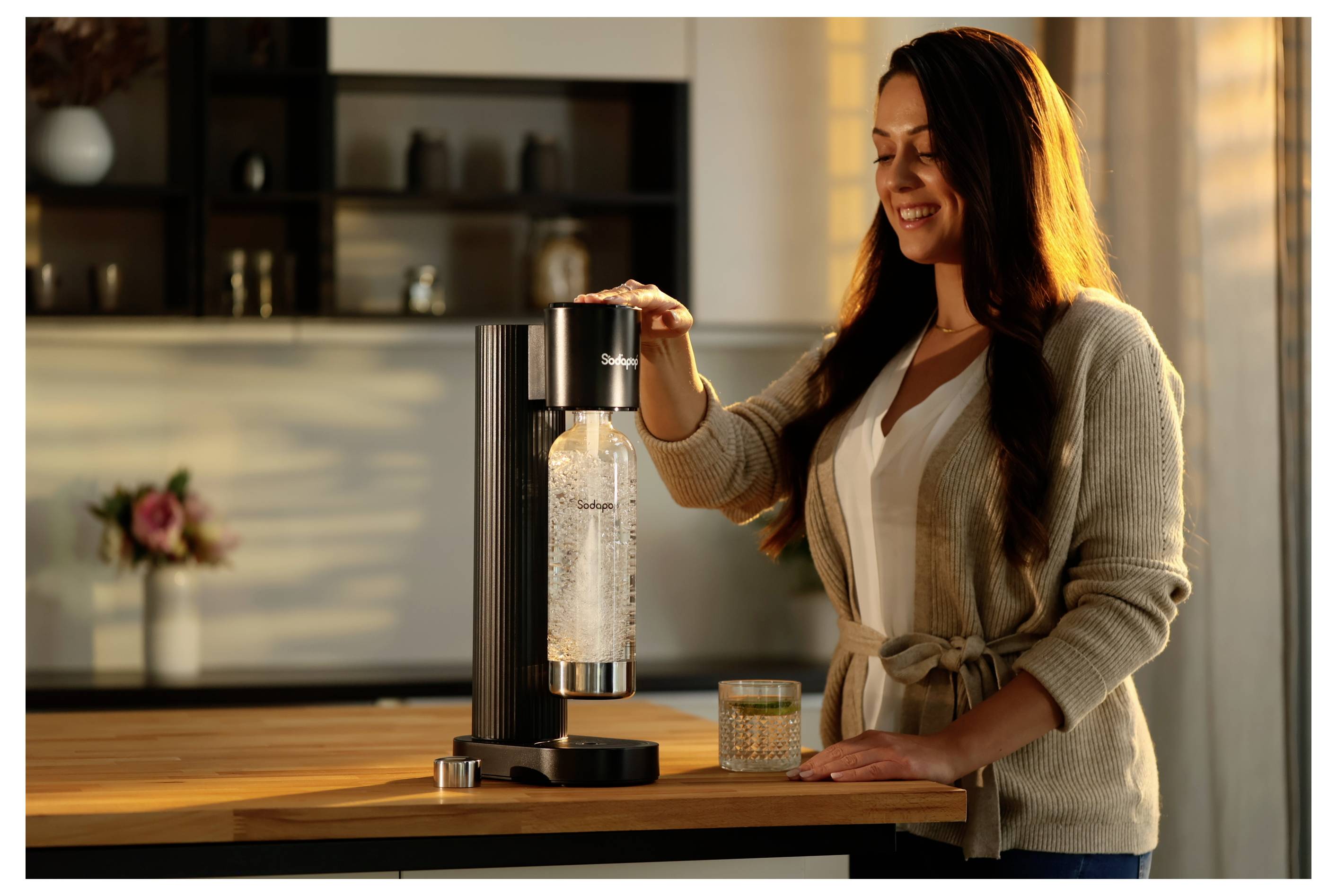 Eine Frau bedient einen Soda-Maker auf einer Küchenablage und lächelt, während sie eine Wasserflasche mit Kohlensäure versetzt. Sonnenlicht fällt durch ein Fenster.